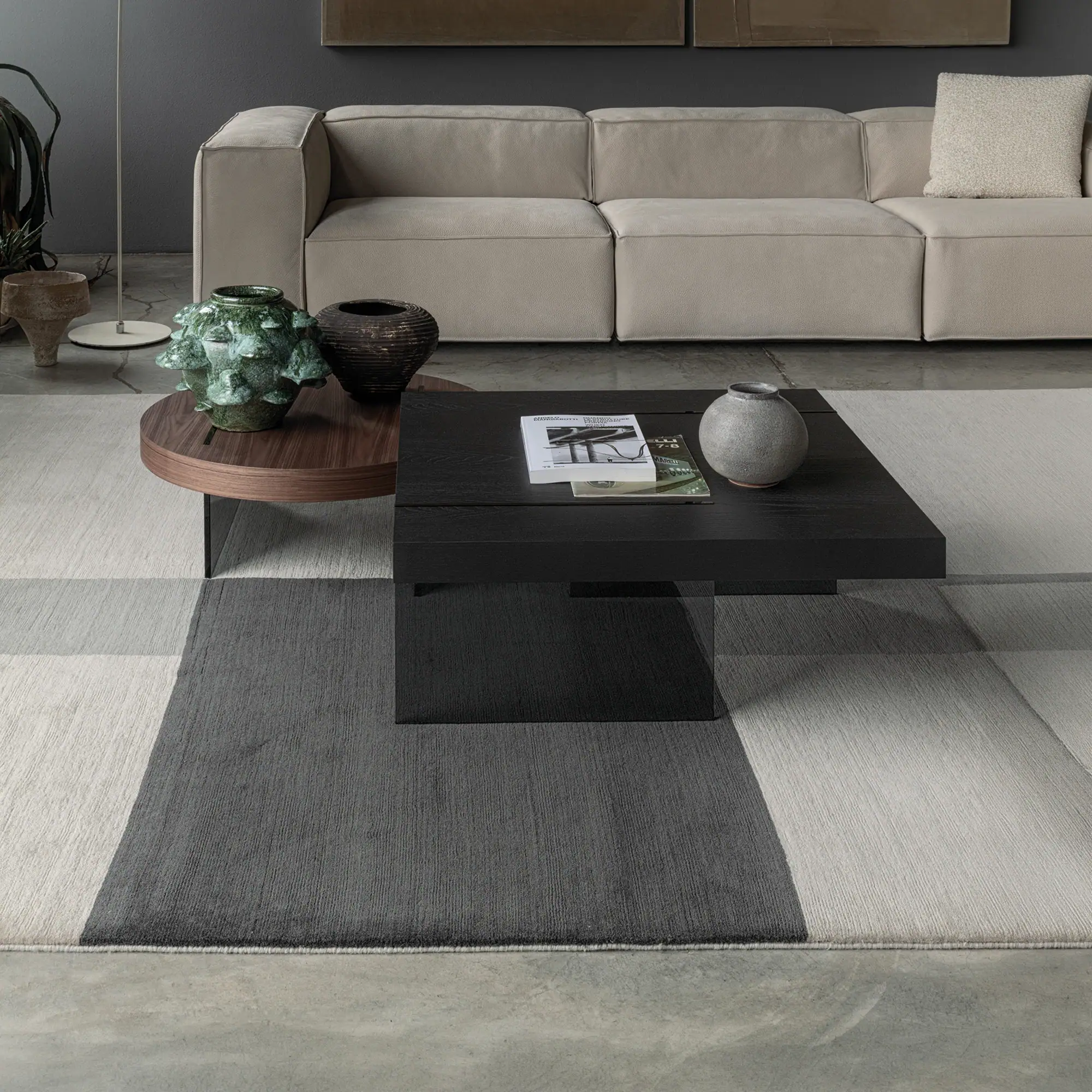 A modern living room with a beige modular sofa, dark coffee tables, and a geometric patterned rug on a concrete floor.