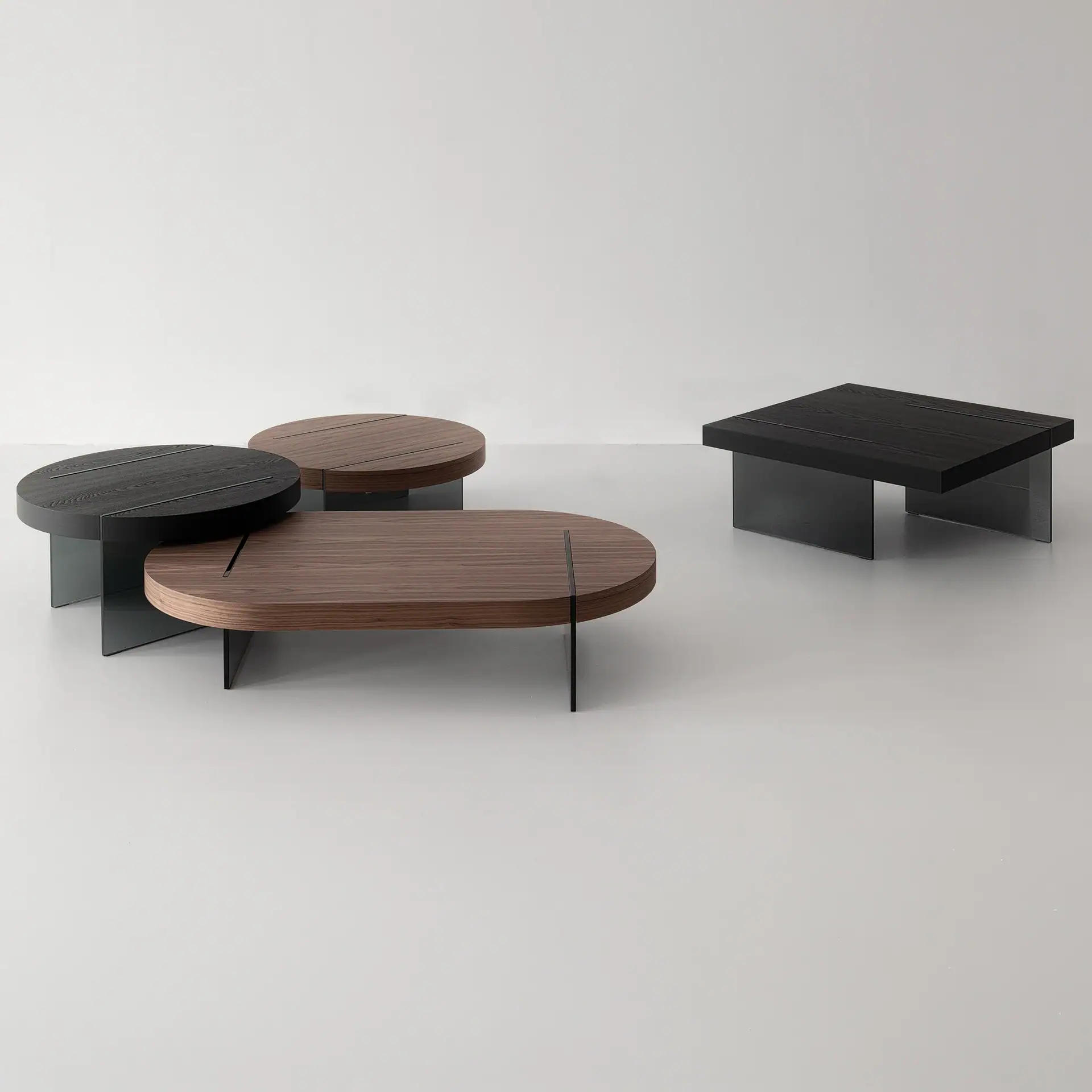 Four modern coffee tables with wooden tops and clear glass legs. Two round, one oval, and one rectangular table are shown, in black and dark brown wood finishes.