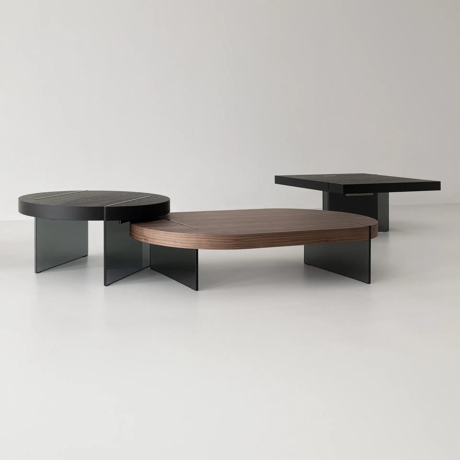 Three modern coffee tables on a light floor against a white wall. One is round with a dark wood top, one is oval with a brown wood top, and one is square with a dark wood top. All have dark glass bases and metal inlays.