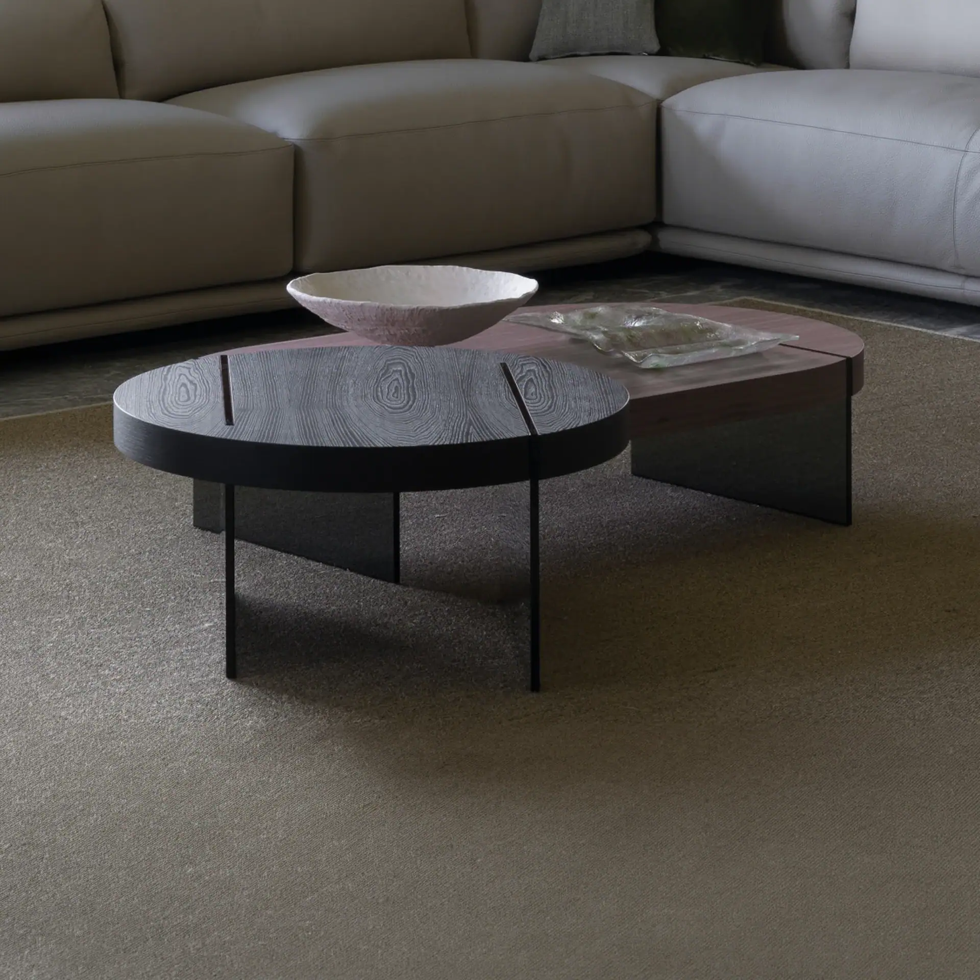 Two modern coffee tables in a living room. The front table is dark wood with a circular top, and the back table is reddish-brown with a textured bowl and glass dish. A light-colored leather sofa is in the background.