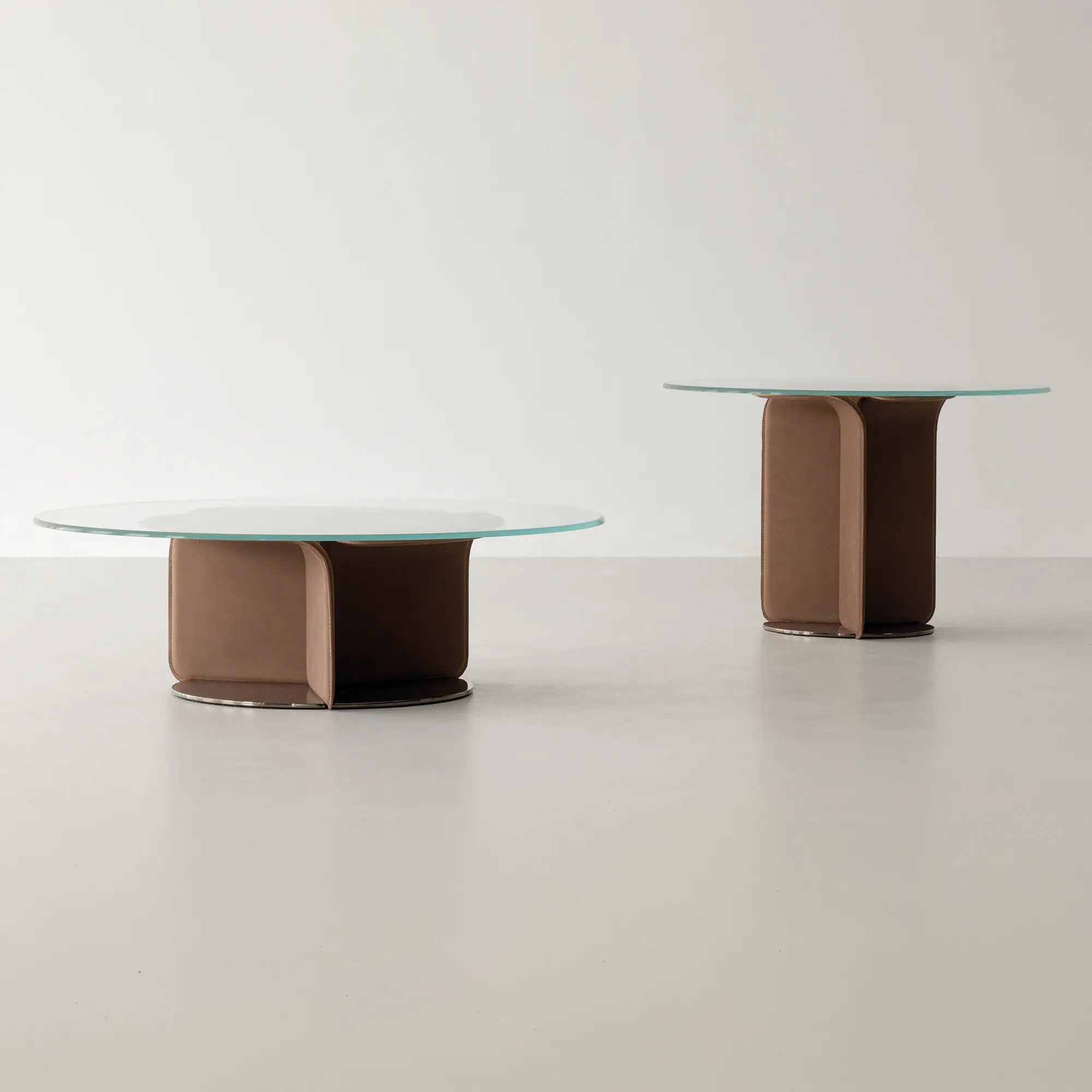 Two modern tables with round glass tops and brown sculptural bases. One is a low coffee table, the other a taller side table.