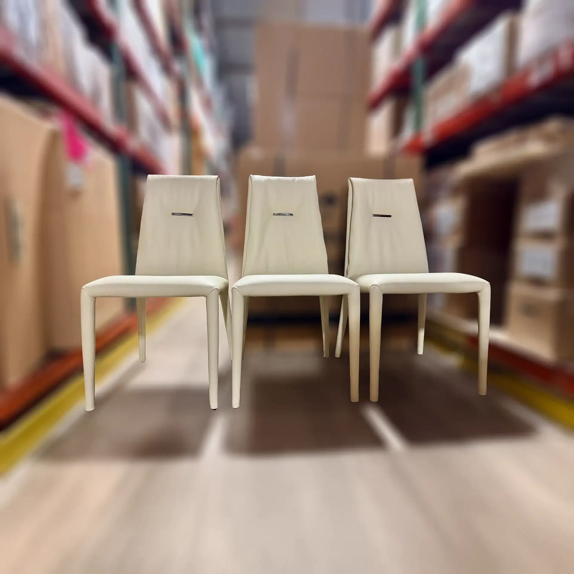 Three identical off-white upholstered chairs with metallic accents on their backrests, arranged in a row against a blurred warehouse background.