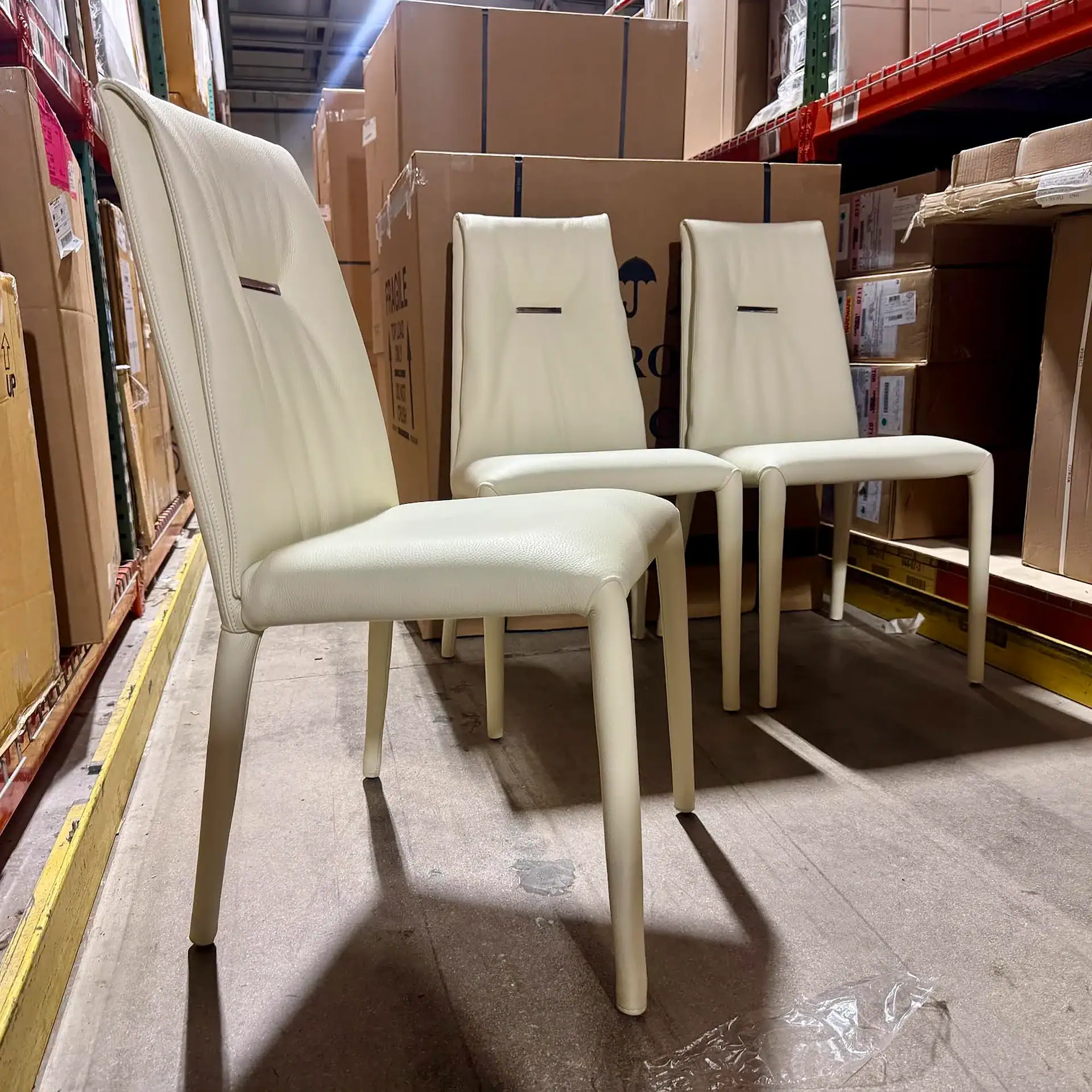Three cream-colored upholstered dining chairs in a warehouse, surrounded by stacked cardboard boxes on metal shelves.