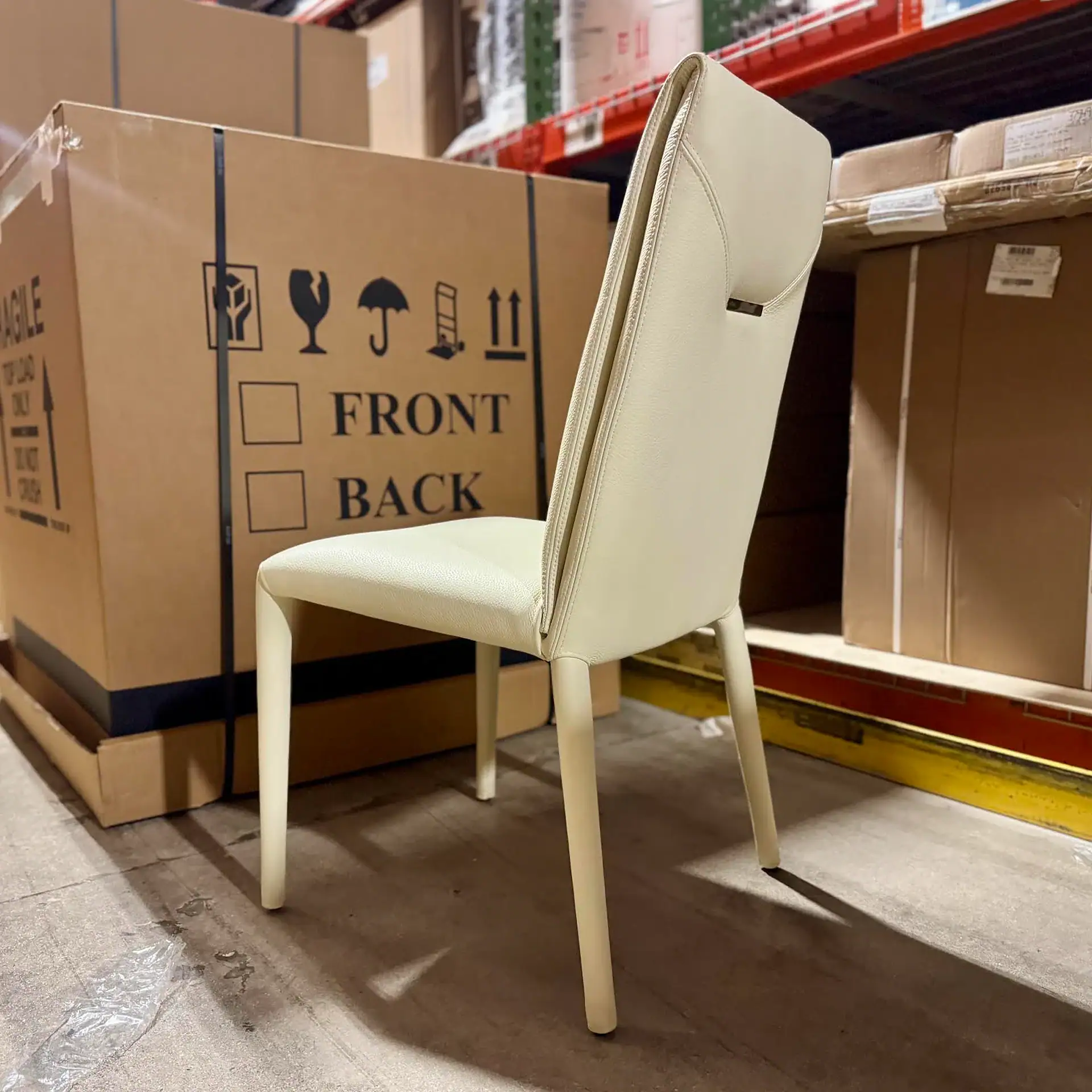 A cream-colored modern dining chair stands in a warehouse, with large cardboard boxes and shelving in the background.