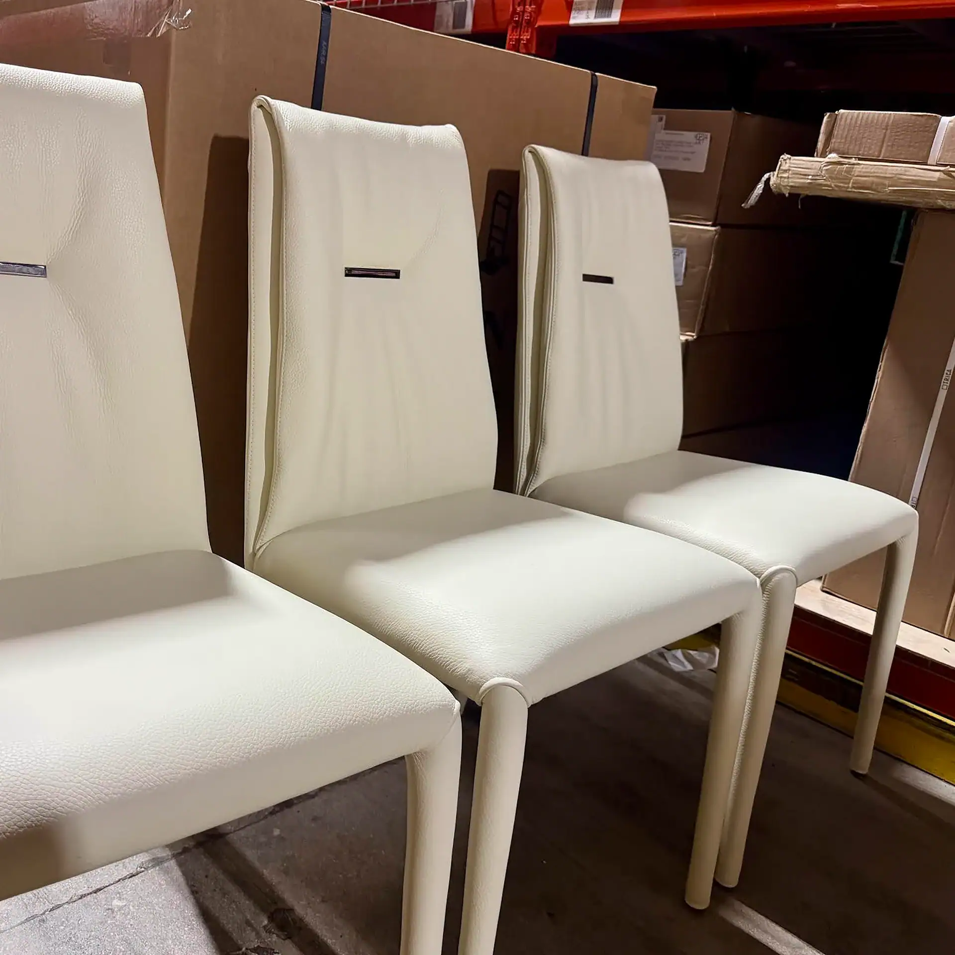 Three cream upholstered dining chairs with high backs and metallic accents, set against a backdrop of cardboard boxes in a warehouse.
