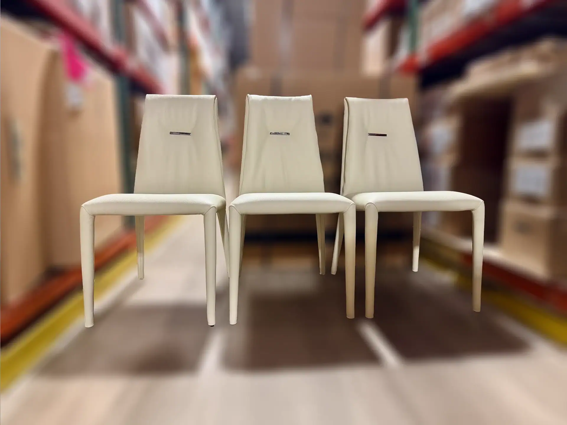 Three cream-colored chairs with metallic accents on their backrests, lined up against a blurred warehouse background.