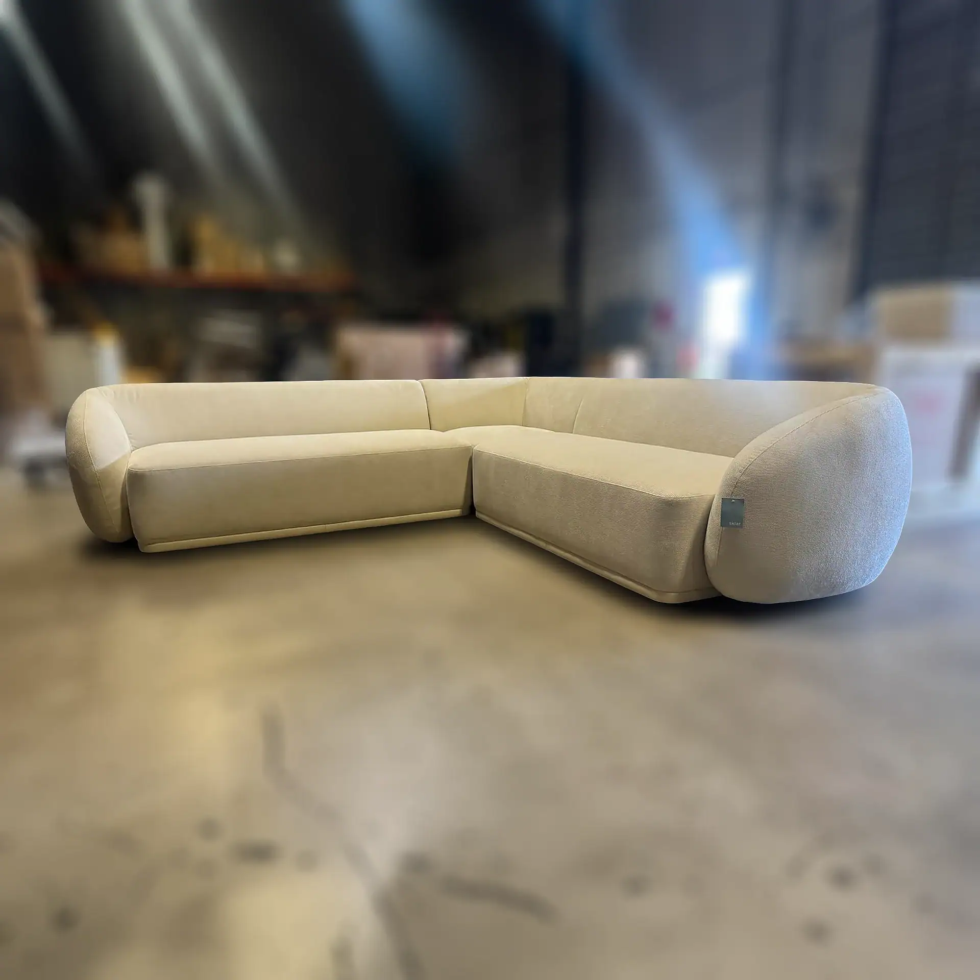 A cream-colored L-shaped sectional sofa with rounded armrests and a 'sklar' tag on the right arm, on a concrete floor with a blurred warehouse background.