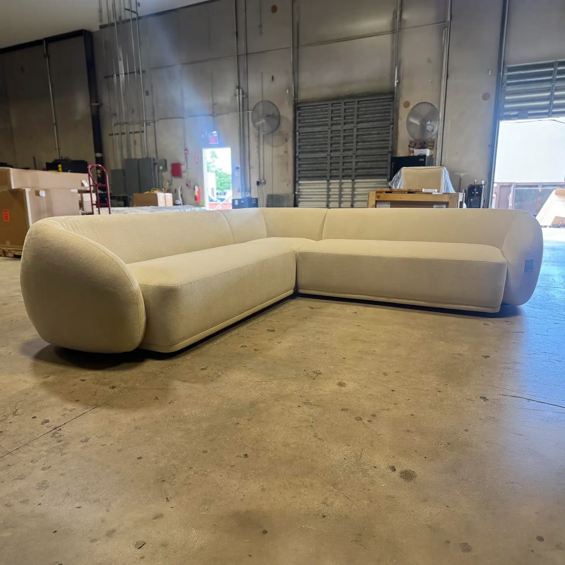 A light cream L-shaped sectional sofa with rounded ends sits on a concrete floor in a warehouse.