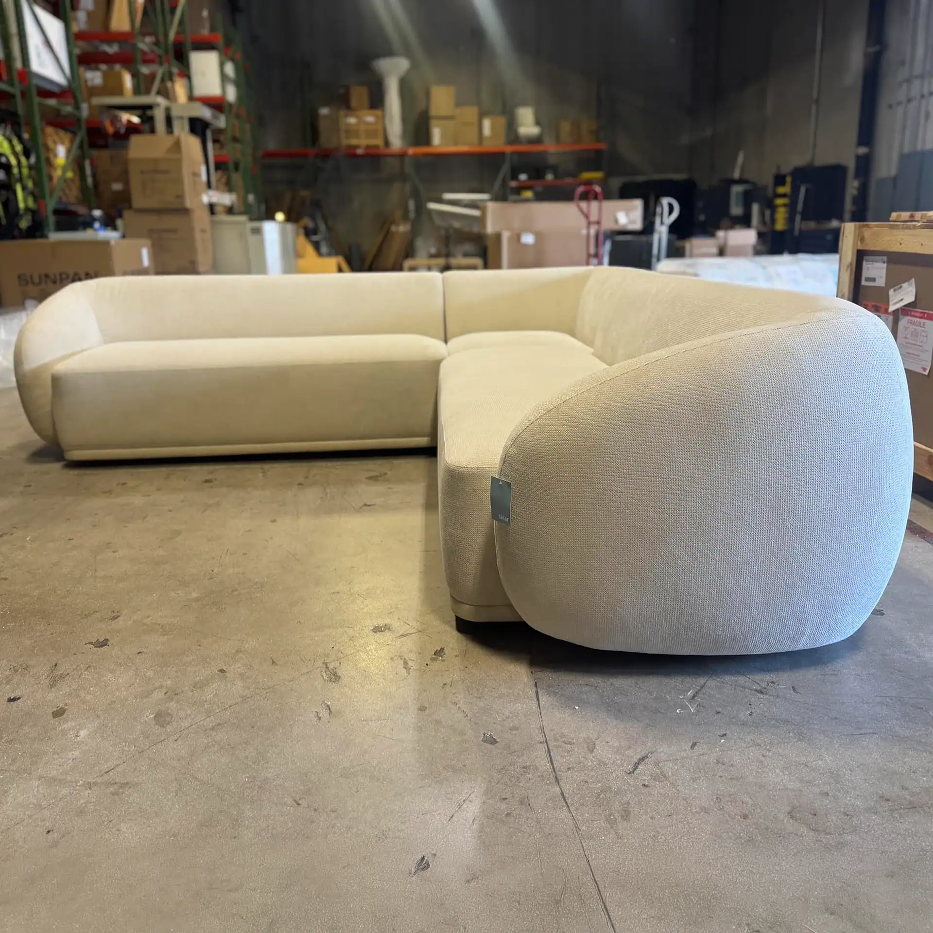 A large, L-shaped cream sectional sofa with rounded arms sits on a concrete floor in a warehouse setting, surrounded by shelves of boxes.