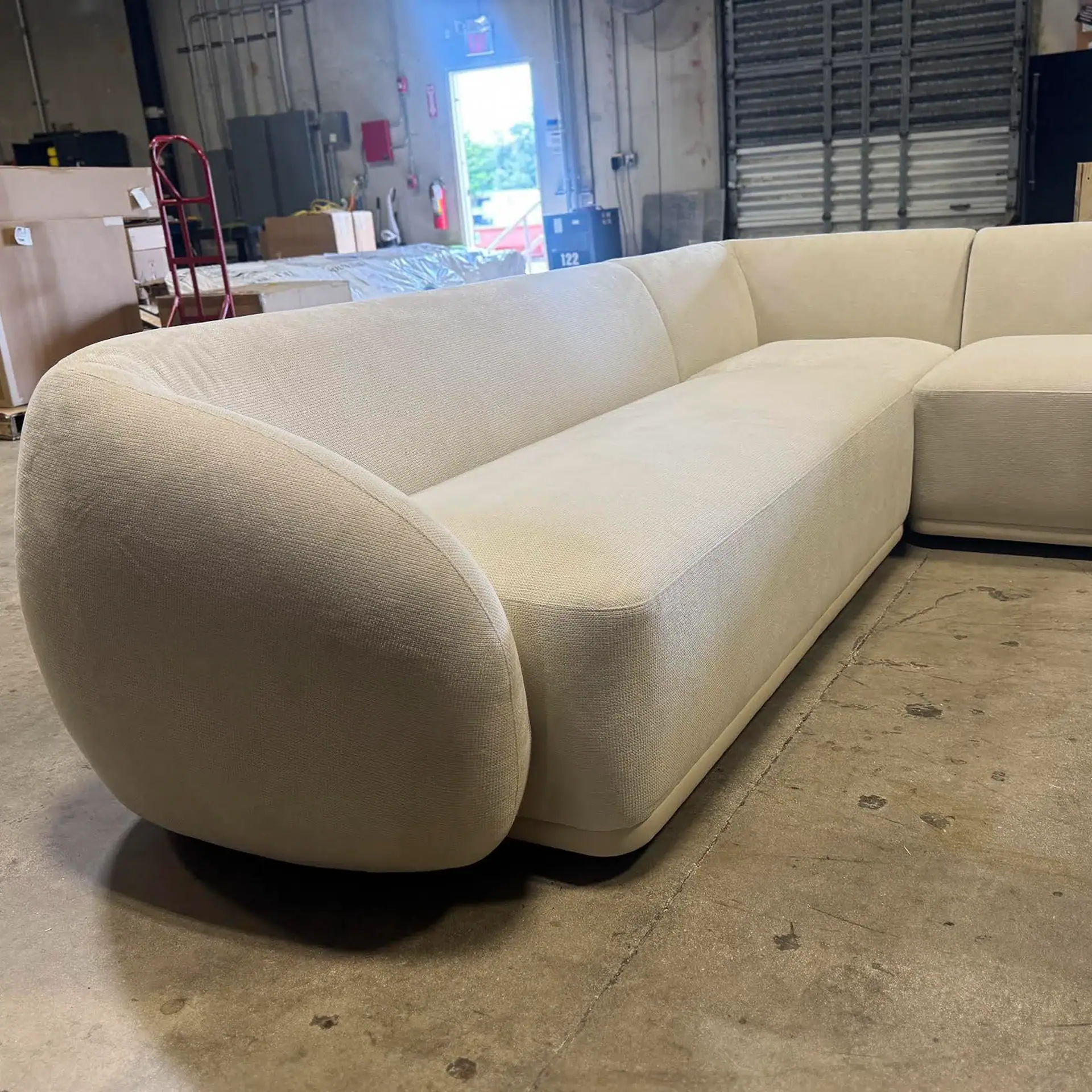 A large, off-white sectional sofa with rounded edges sits on a concrete floor in a warehouse, surrounded by industrial equipment and boxes.