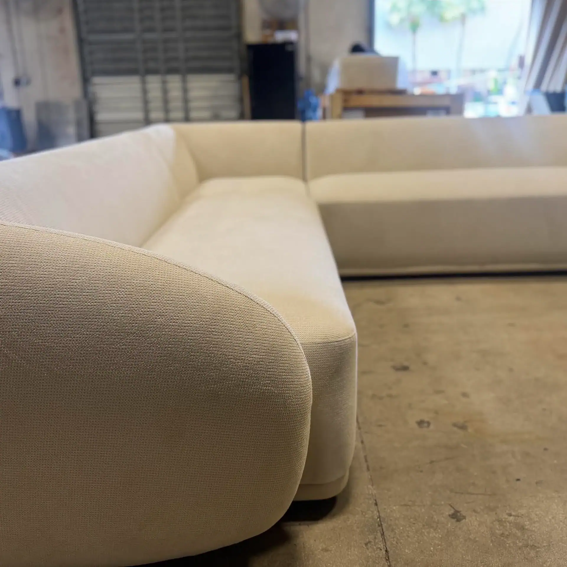 A cream-colored, L-shaped sectional sofa with rounded edges and textured fabric, set on a concrete floor in an industrial space with a blurry background showing a window and shelving.