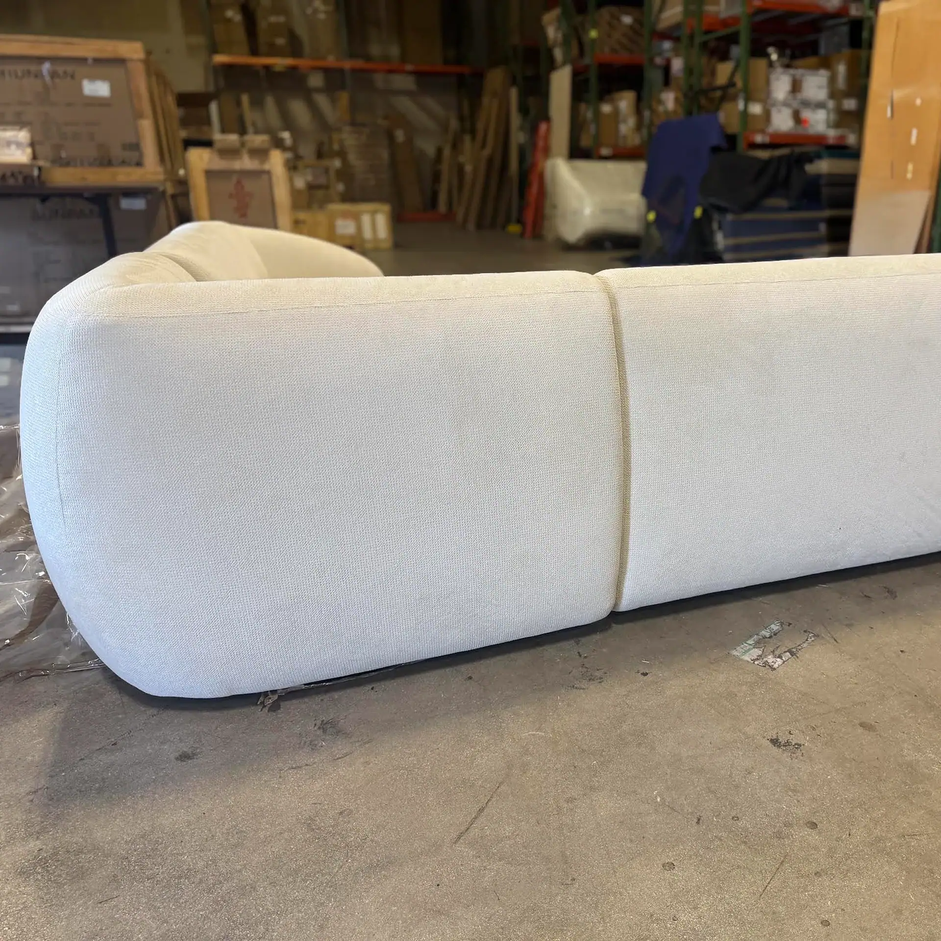 A light-colored modular sofa with a rounded armrest, secured with a rope, sitting on a concrete floor in a warehouse.