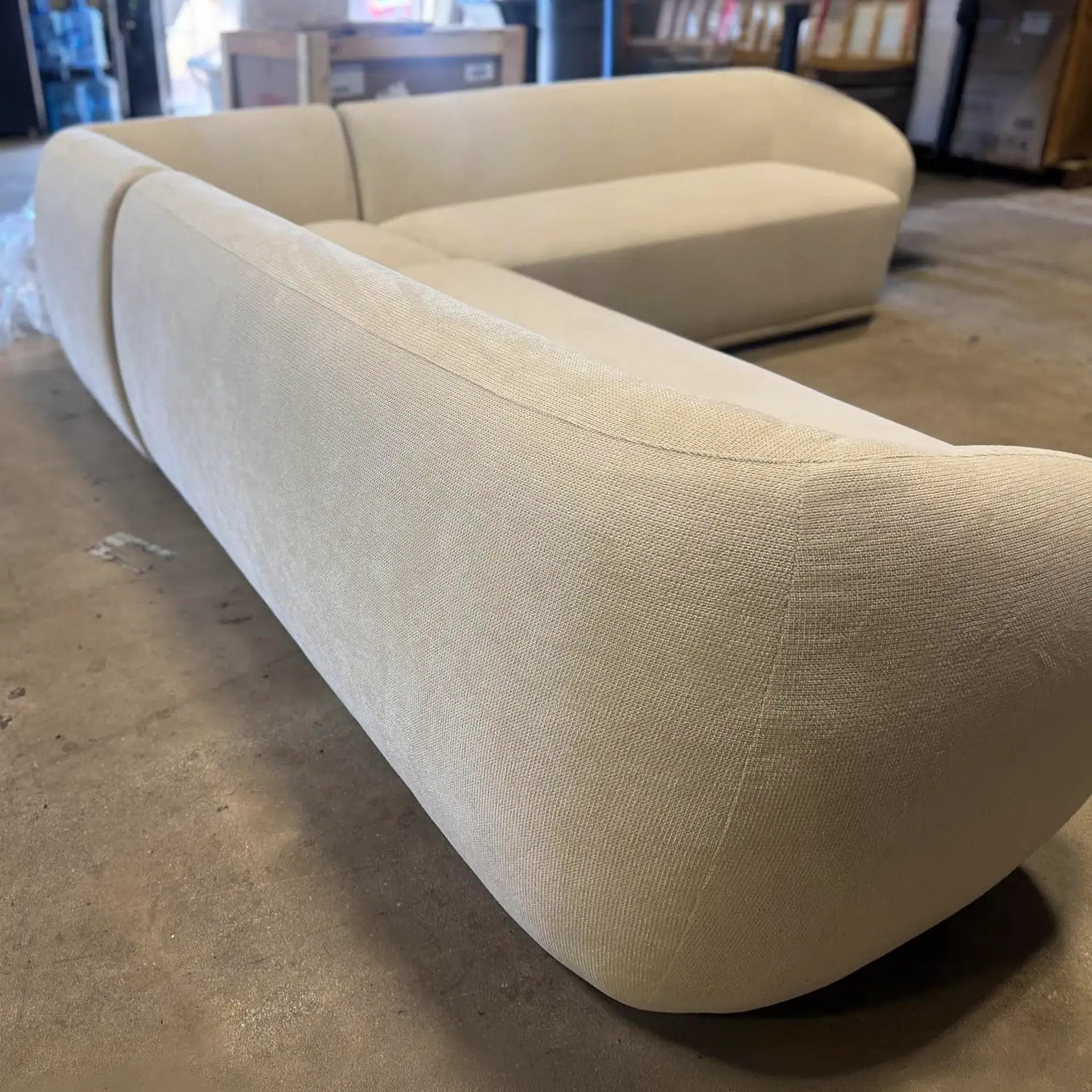 A large, light beige L-shaped sectional sofa with rounded edges sits on a concrete floor in a warehouse.