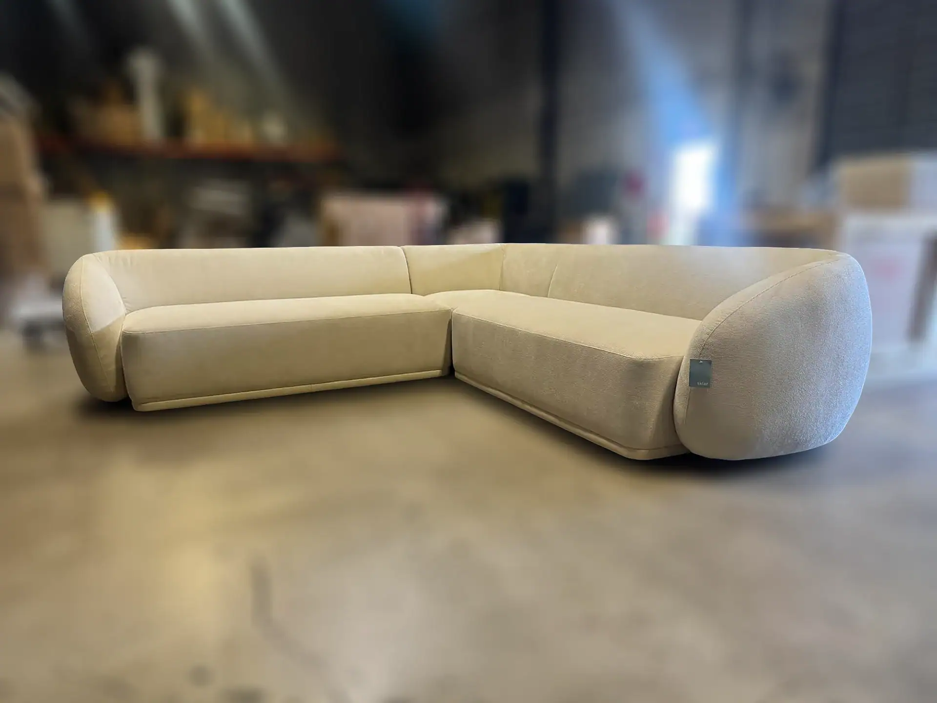 A light beige, L-shaped sectional sofa with rounded edges and a textured fabric, displayed on a smooth floor in a blurred warehouse-like setting.
