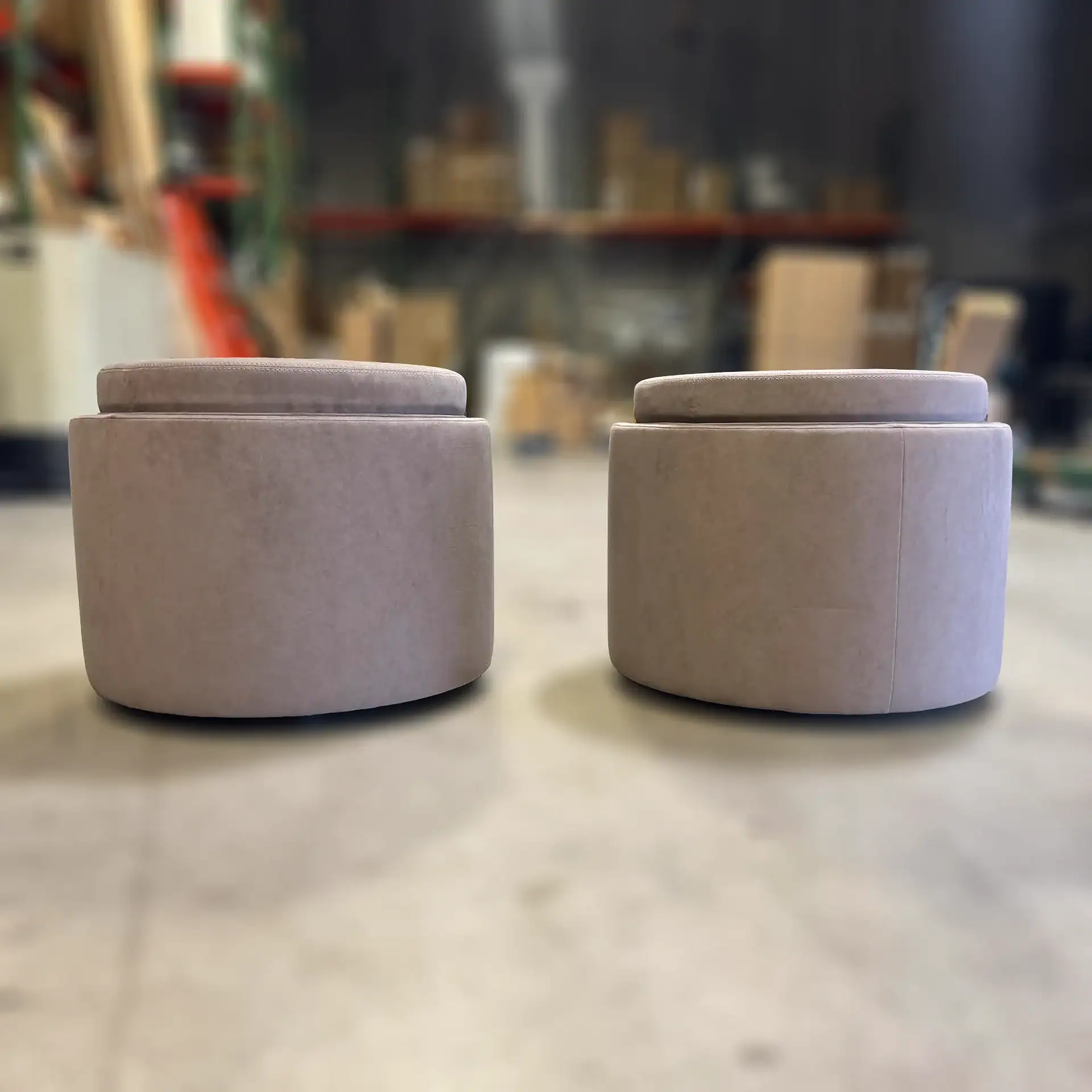 Two light-colored, round upholstered ottomans with stitched tops, shown on a light floor with a blurred warehouse background.
