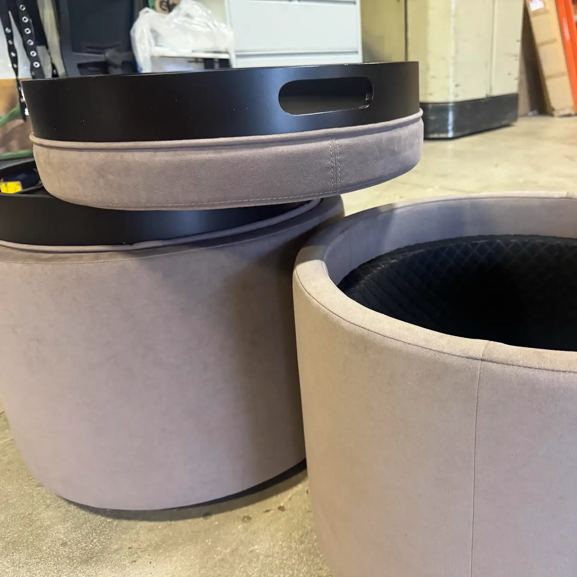 Three round, light grey upholstered ottomans with black tray lids. Two are stacked, and one is open, revealing a black textured interior.