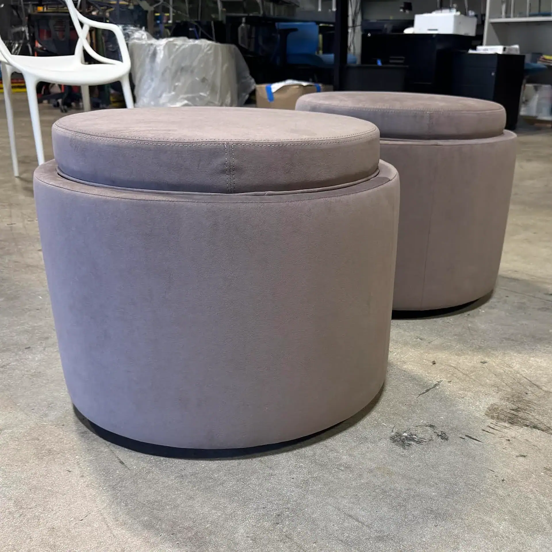 Two round, grayish-purple upholstered ottomans with removable tops on a concrete floor in an industrial setting.
