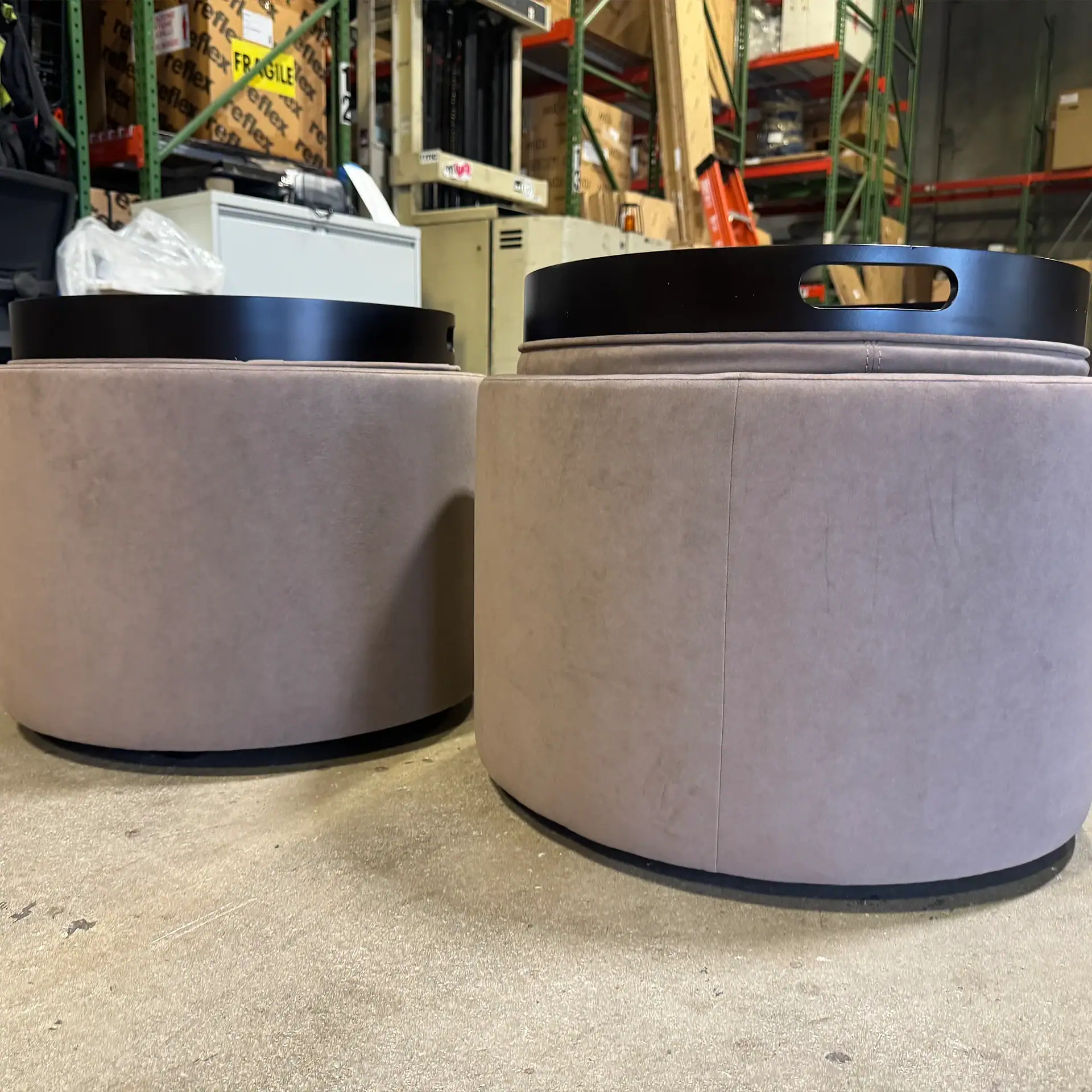 Two round, light brown upholstered ottomans with black trays on top, sitting on a concrete floor in a warehouse.