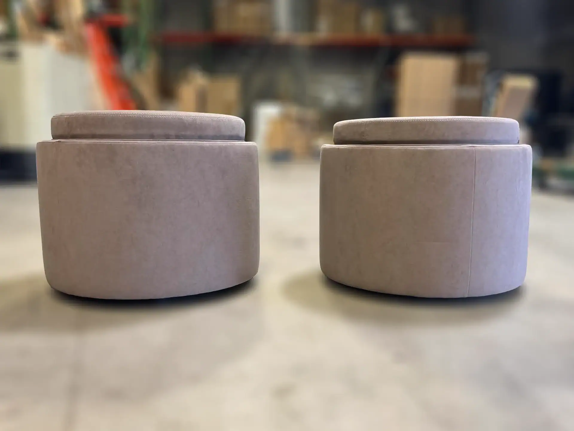 Two light beige round upholstered ottomans with padded tops sit on a light floor, with a blurred warehouse in the background.