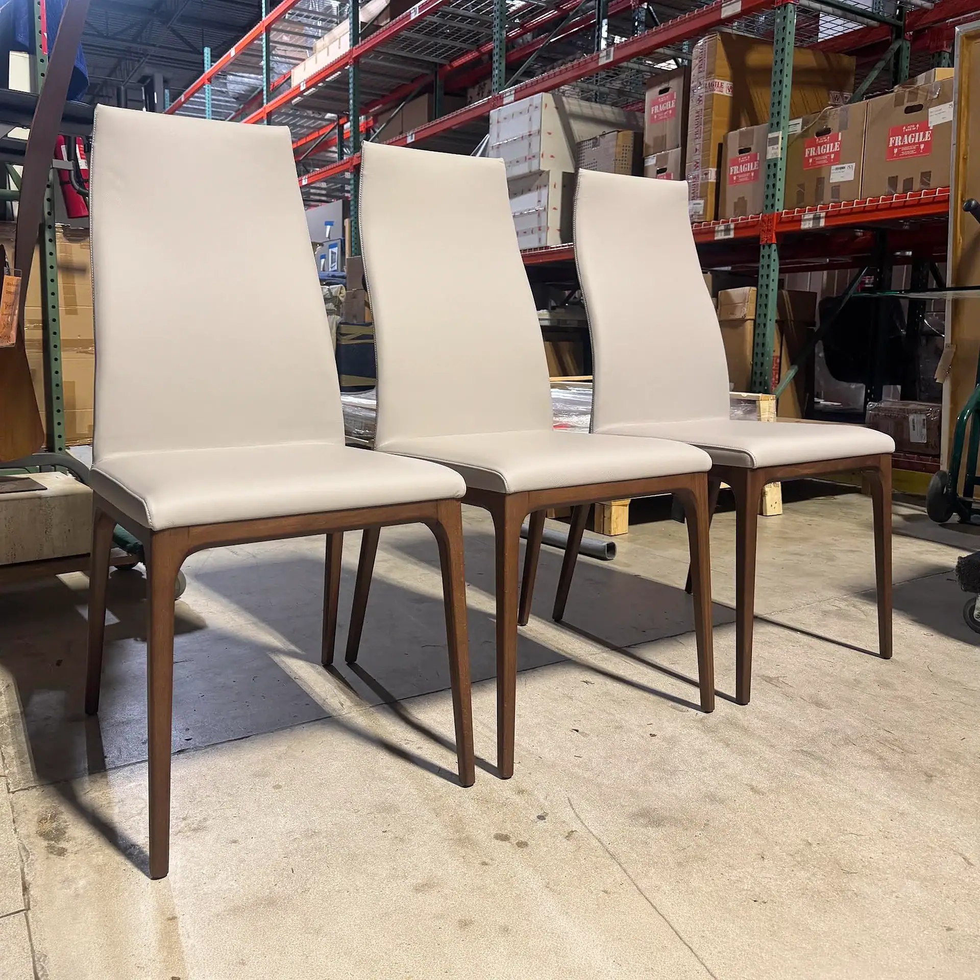 Three light beige upholstered dining chairs with dark wood frames lined up in a warehouse setting.