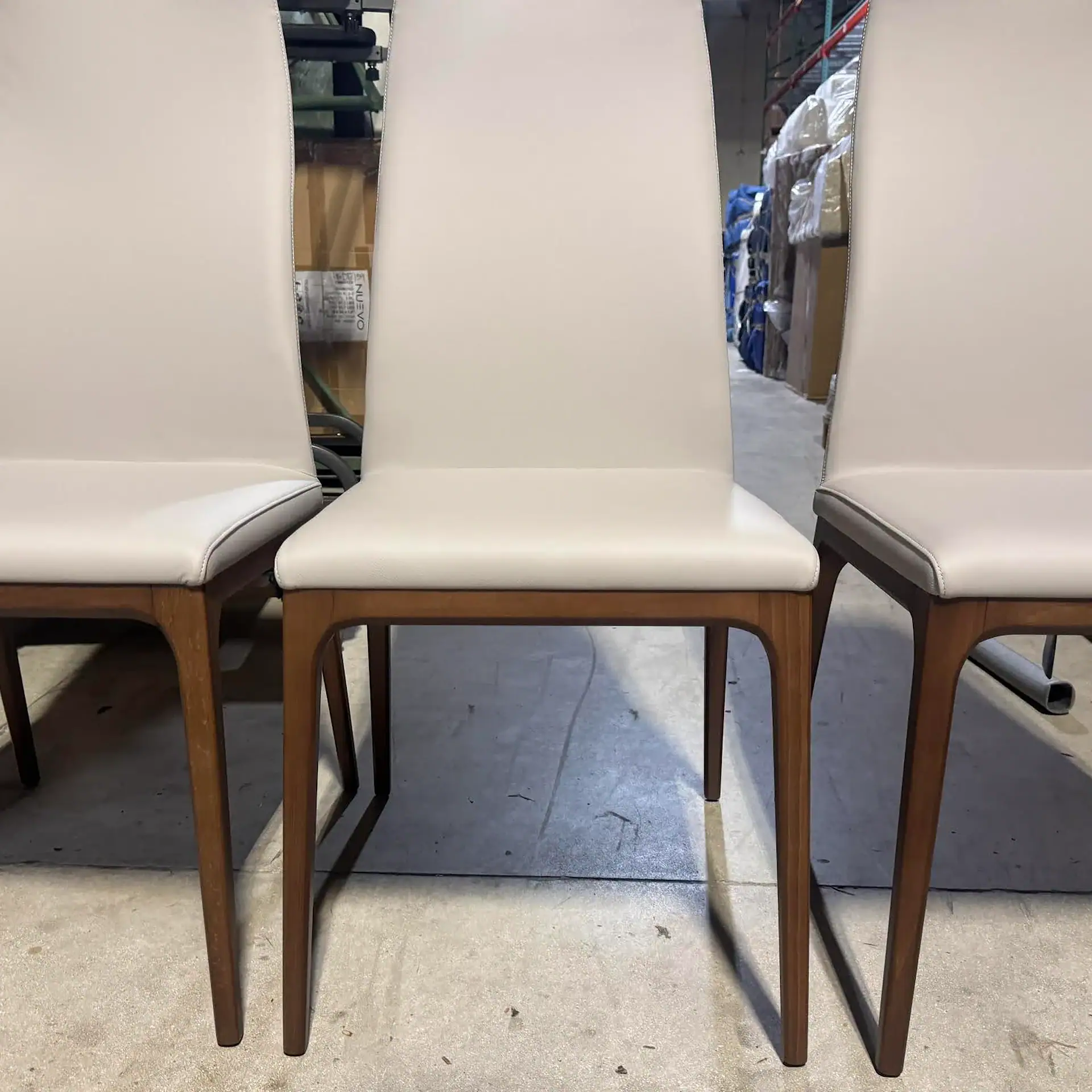Three light beige upholstered dining chairs with dark wooden legs, lined up in a warehouse setting.