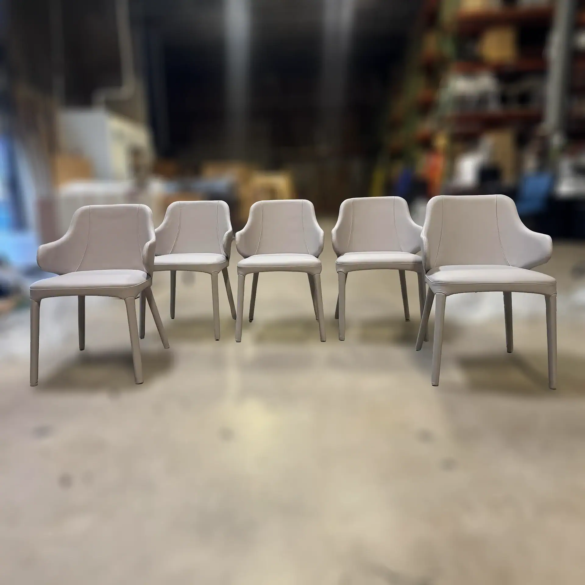 Five light gray upholstered chairs with armrests lined up on a light floor in a blurred industrial setting.