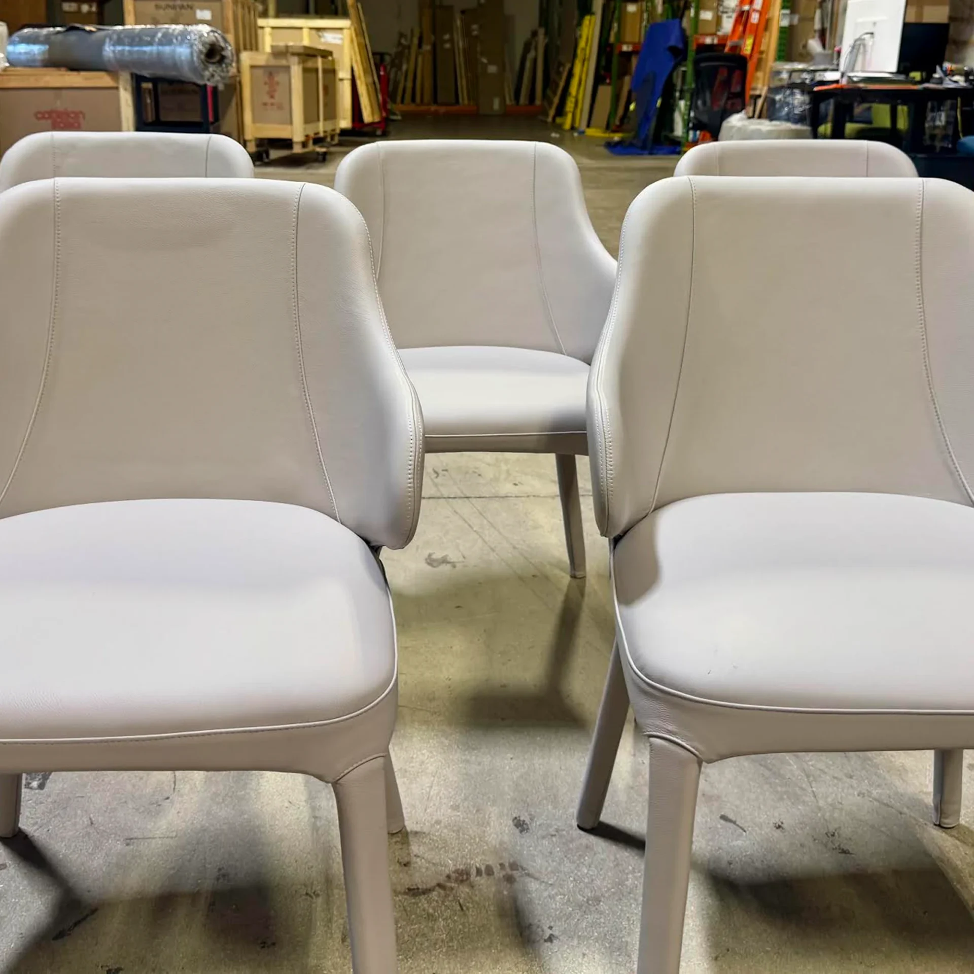 Five modern light gray upholstered chairs with rounded backs and integrated armrests, arranged on a concrete floor in a warehouse.