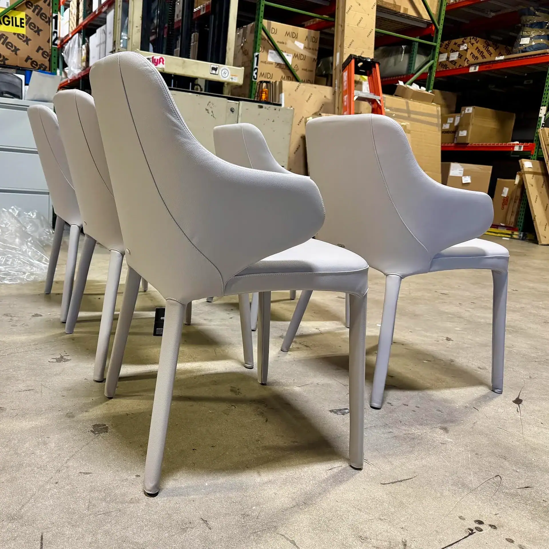 Four light grey upholstered chairs in a warehouse with shelves of boxes and a forklift in the background.