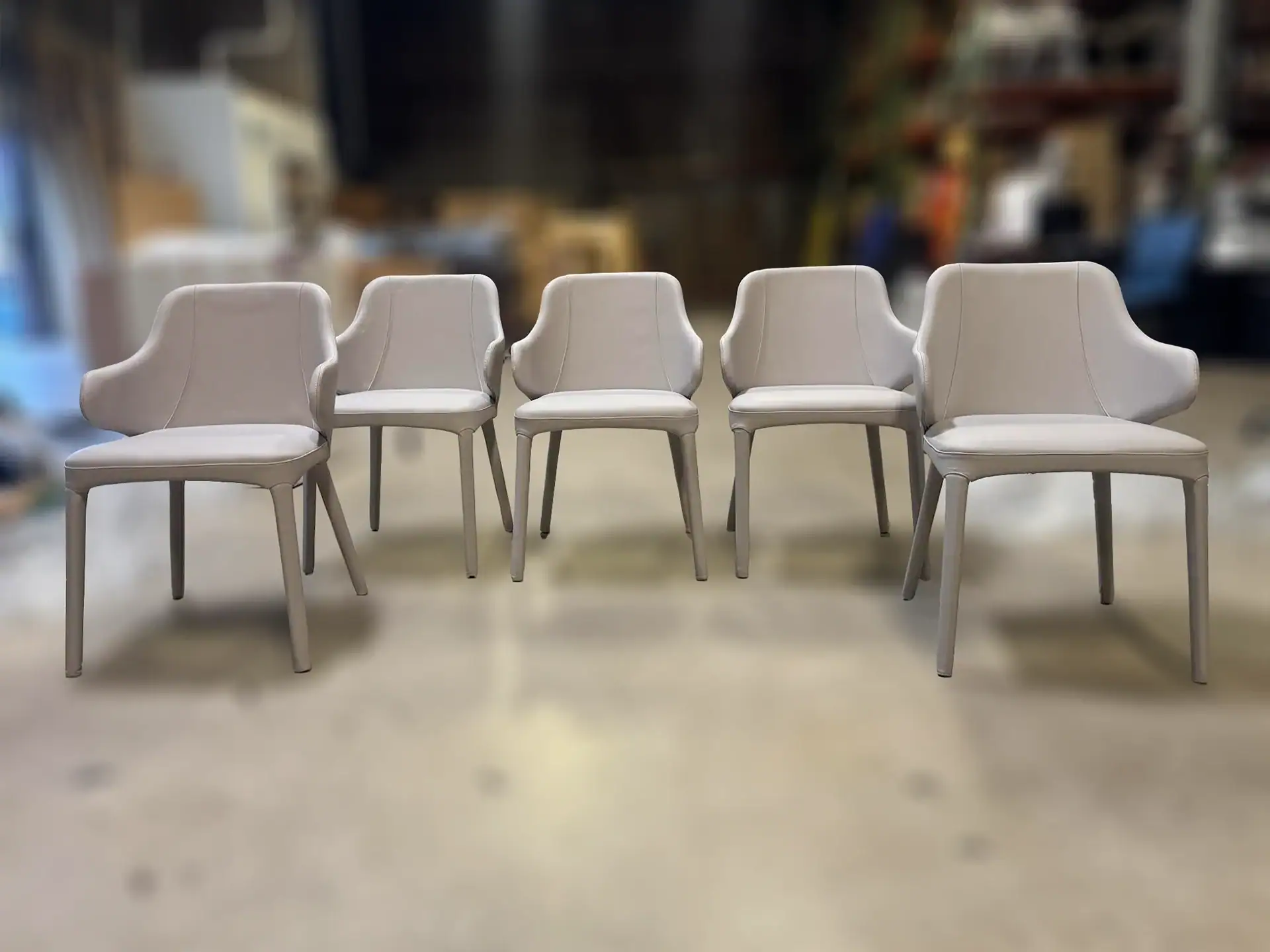 Five identical light grey upholstered chairs with armrests, lined up on a light floor against a blurry background.