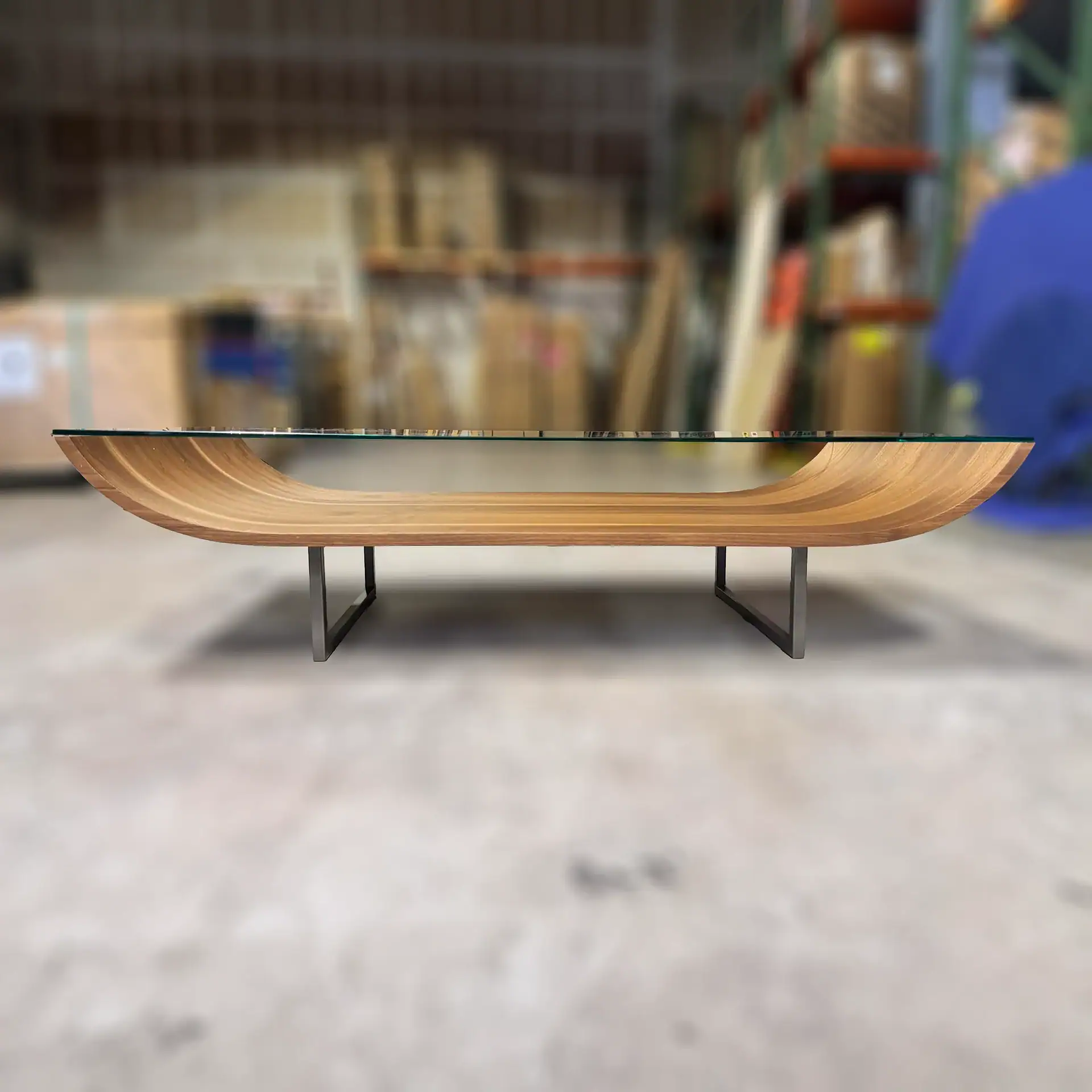 Modern coffee table with a clear glass top and a curved, slatted light wood base on dark metal legs, set against a blurred warehouse background.