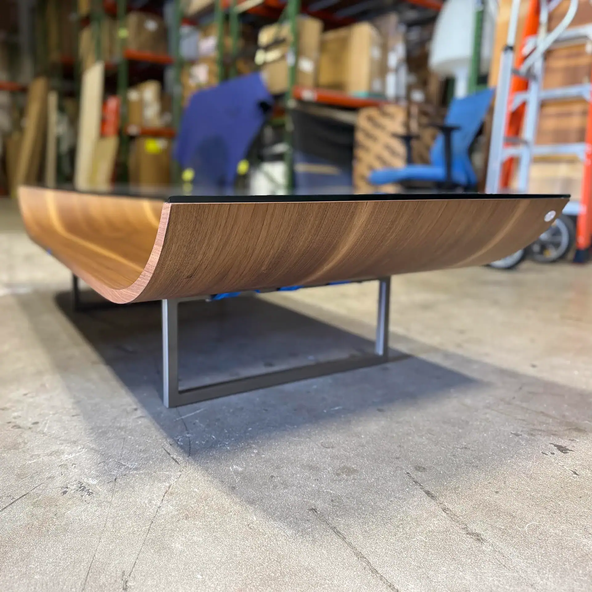 A modern coffee table with a curved wooden base and a black top, set against a blurred warehouse background.