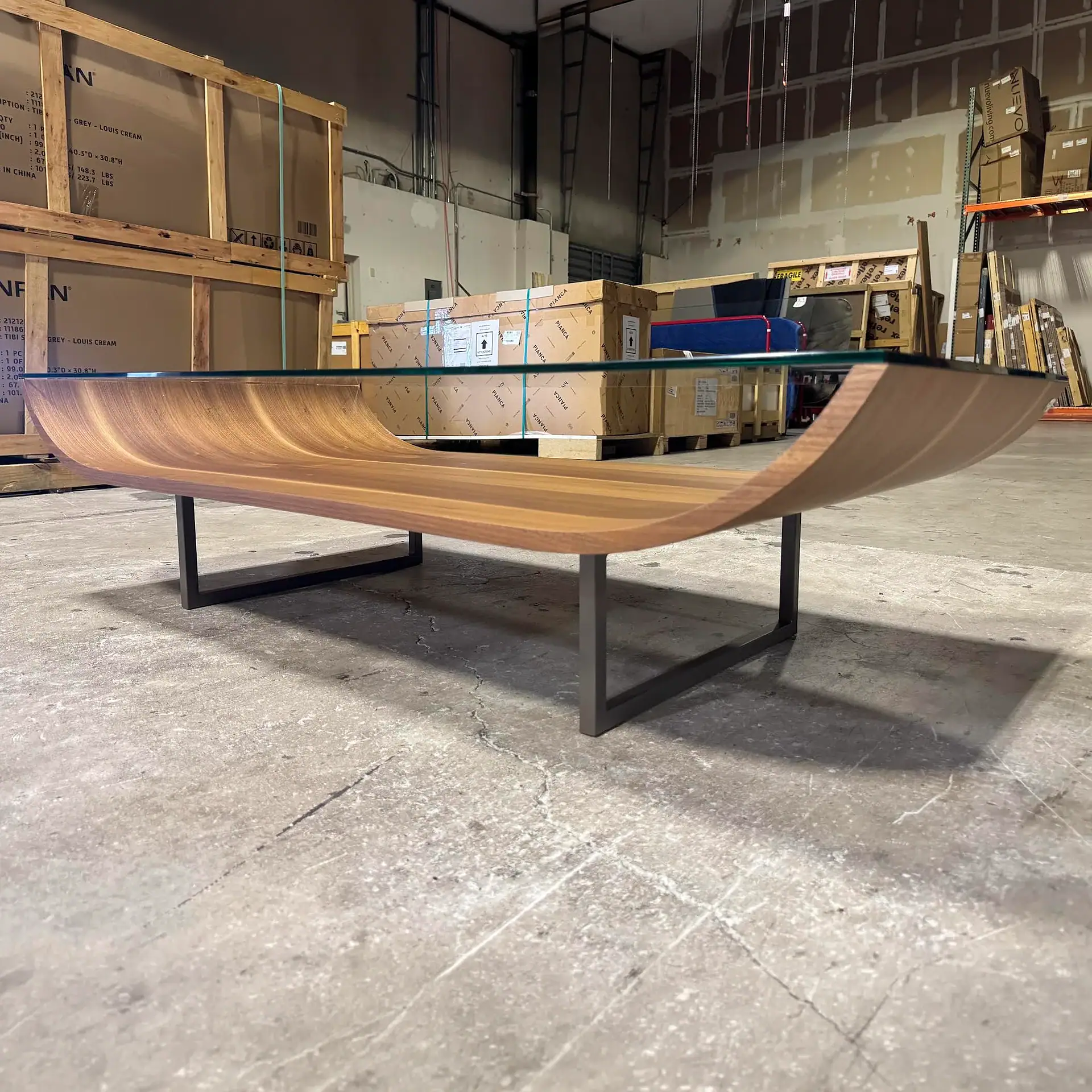 A modern coffee table with a curved wooden base and glass top sits on a concrete floor in a warehouse.
