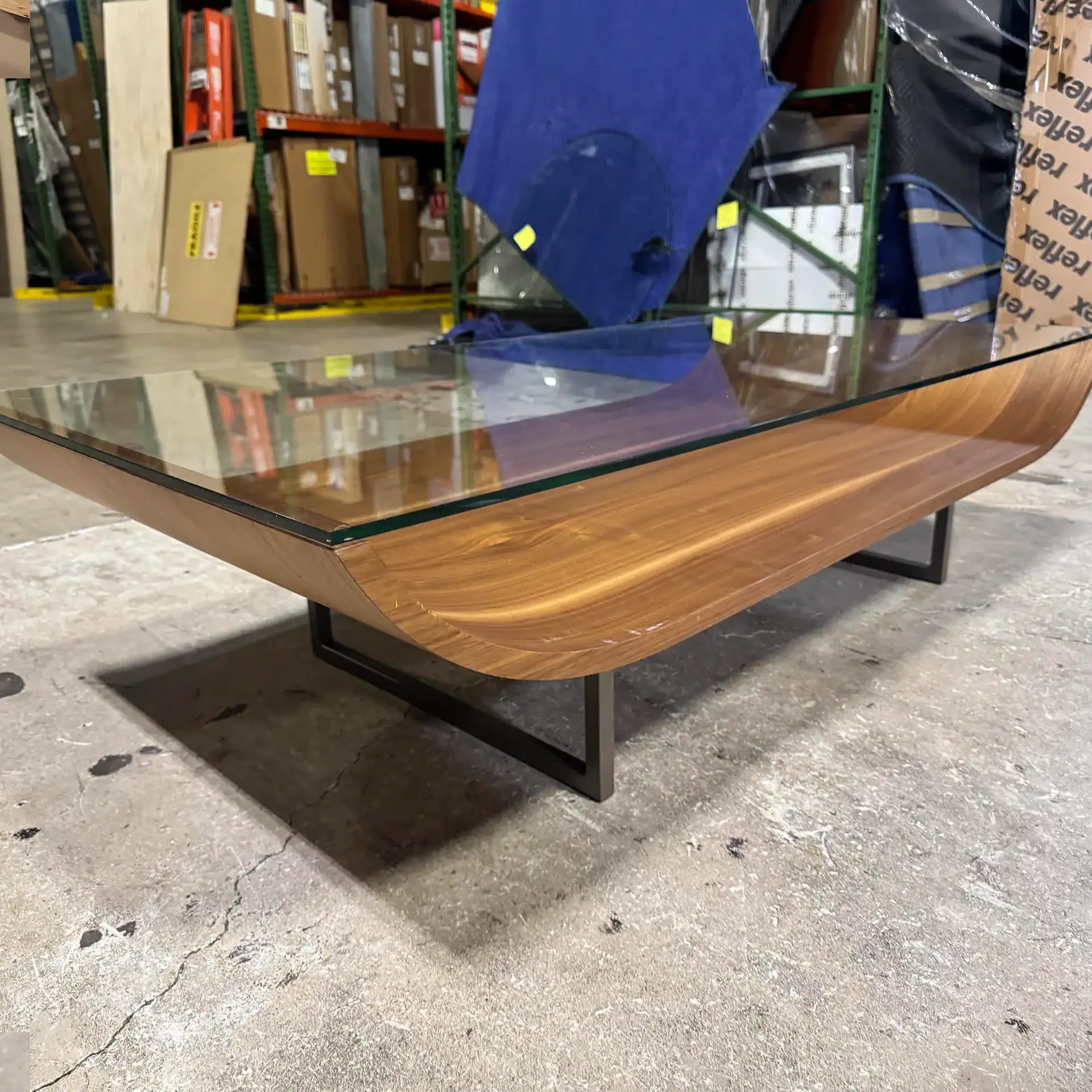 A modern coffee table with a rectangular glass top and a curved, boat-shaped wooden base on dark metal legs, set on a concrete floor in a warehouse.
