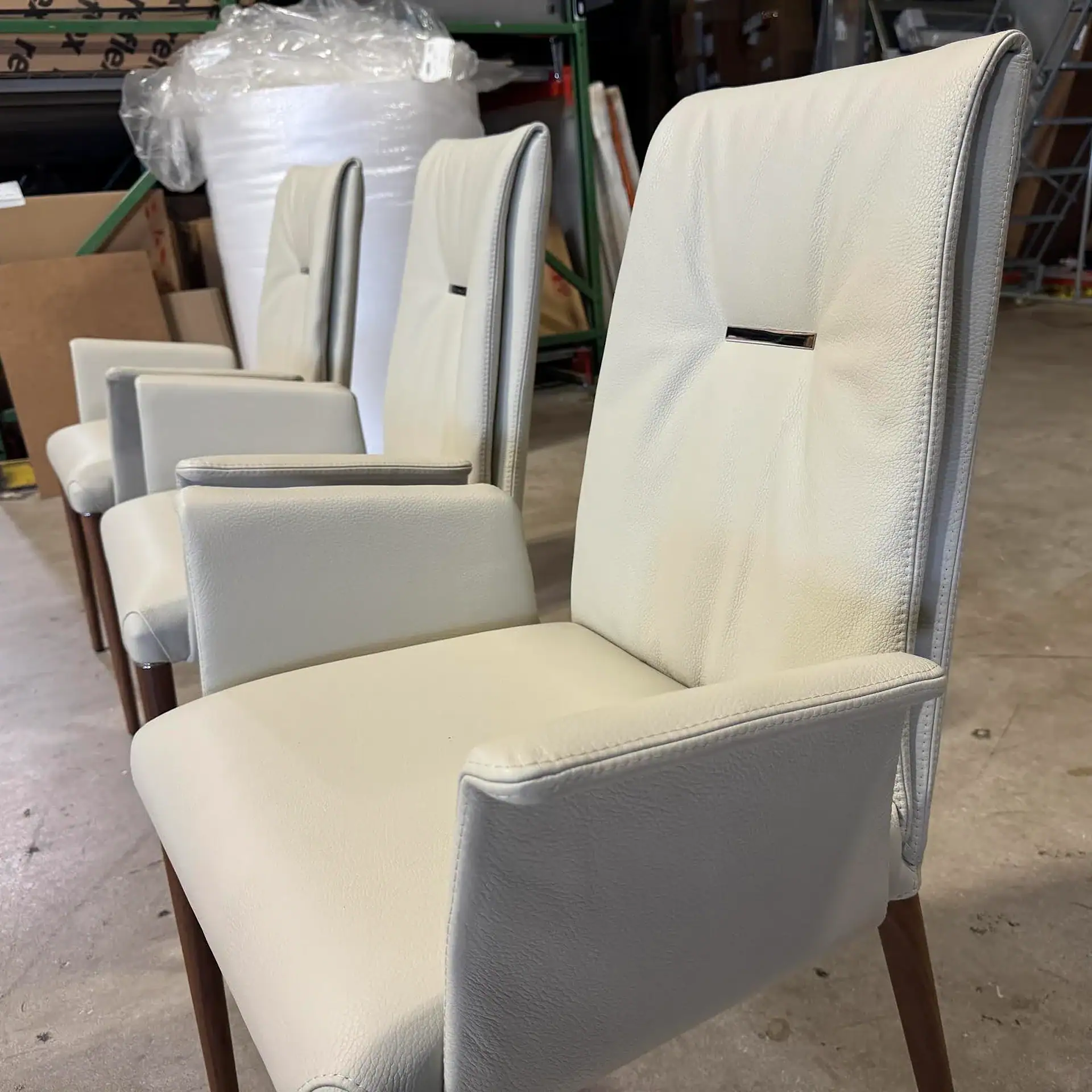 Four cream-colored upholstered dining chairs with armrests and wooden legs, lined up in a row in a warehouse.
