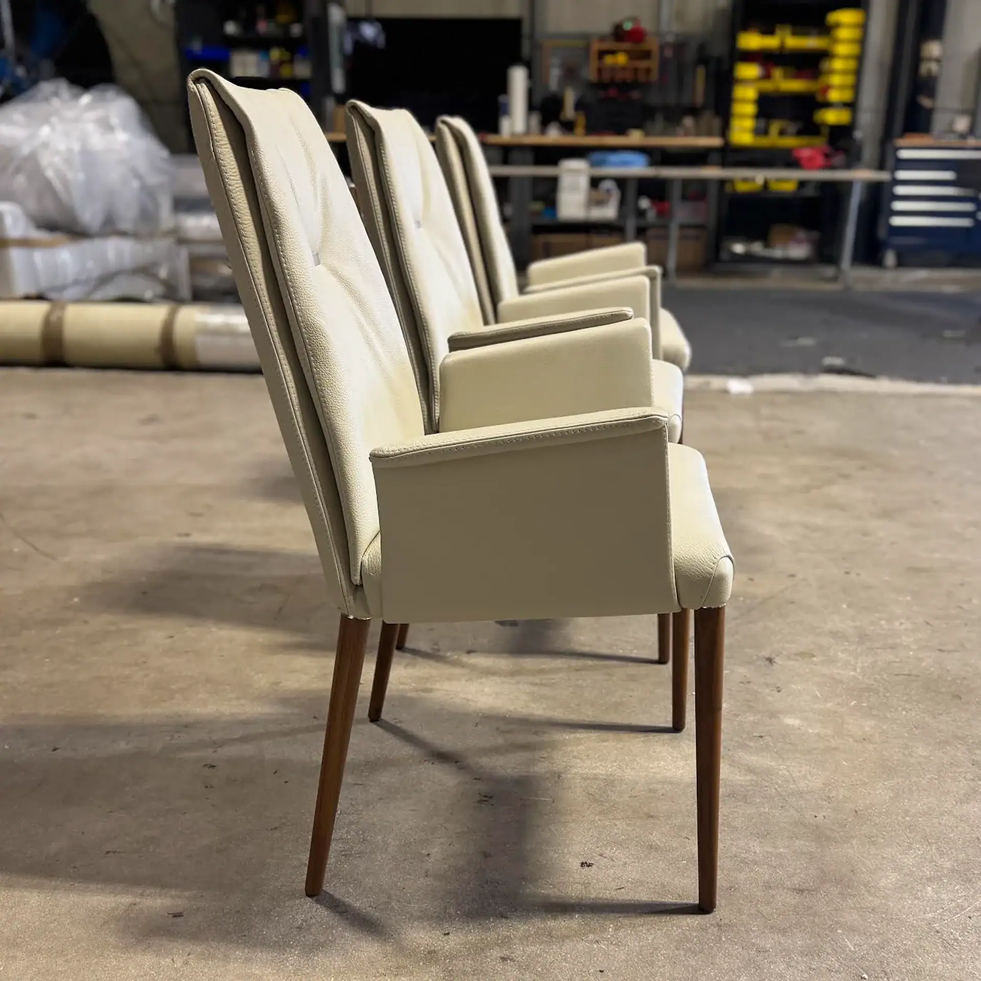 Three cream leather dining chairs with wooden legs in a row on a concrete floor, with a blurred industrial background.