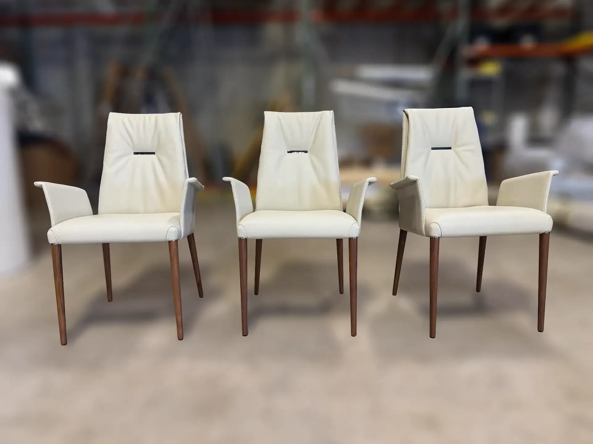 Three identical cream-colored armchairs with dark wooden legs and a black horizontal accent on the backrest, arranged in a row on a light floor.