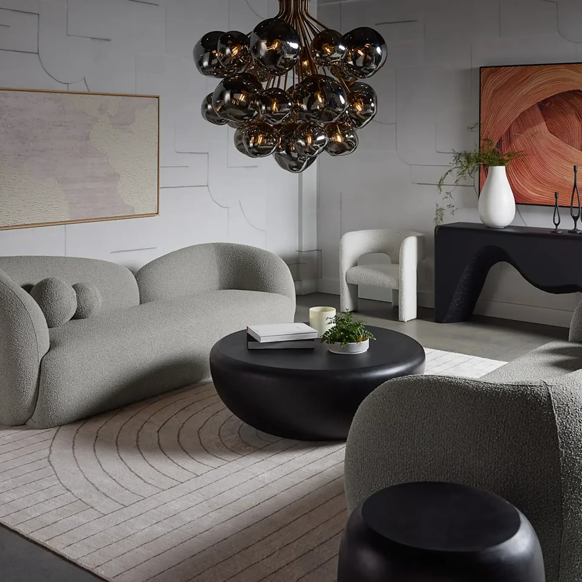 A modern living room with a grey curved sofa, black coffee table, and a chandelier with dark spherical shades. Textured walls and abstract art complete the contemporary look.