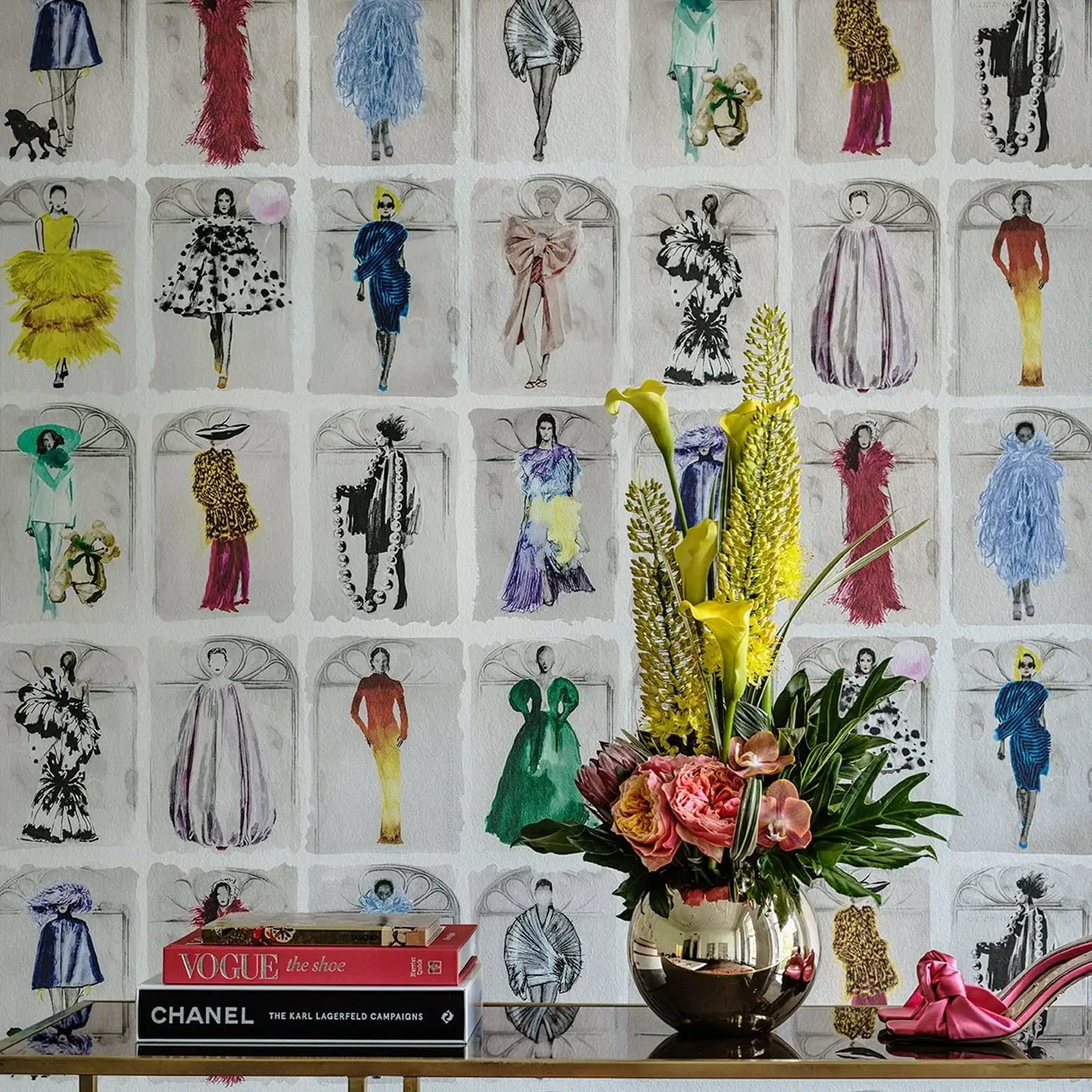 A console table with fashion books, a metallic vase holding yellow and pink flowers, and pink high heels, set against a wallpaper patterned with colorful fashion illustrations.