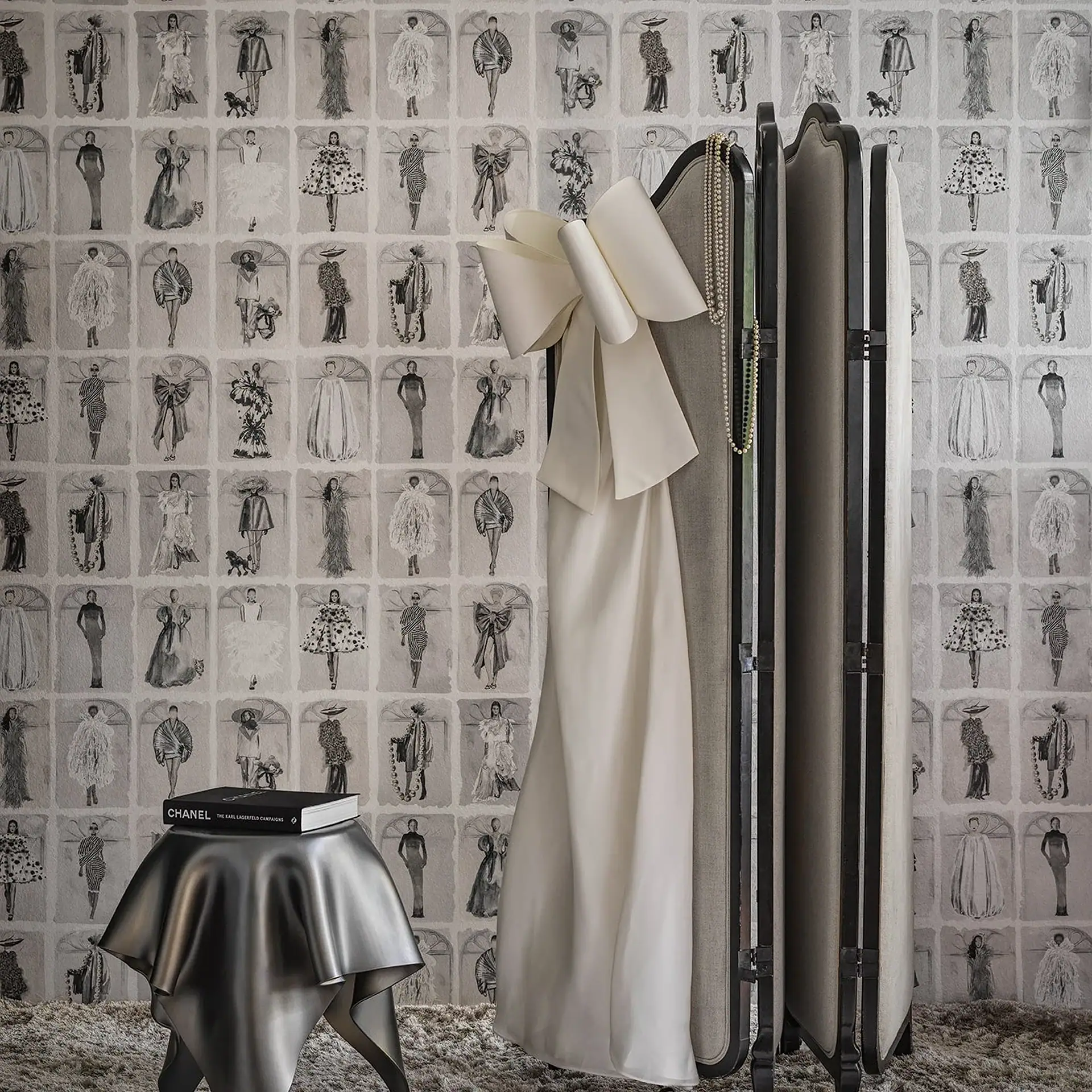 An off-white dress with a large bow hangs on a folding screen in front of a wallpaper patterned with black and white fashion illustrations. A black Chanel book rests on a metallic silver sculptural stool.