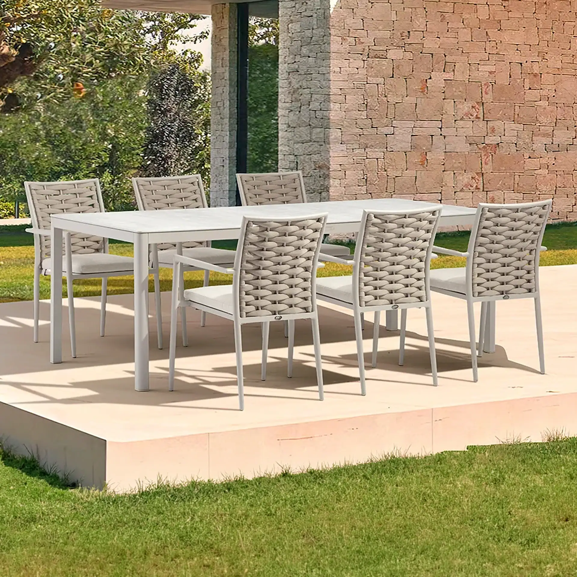A modern outdoor dining set with a light grey table and eight chairs with woven backs and cushions, on a patio surrounded by green grass and a stone wall building.