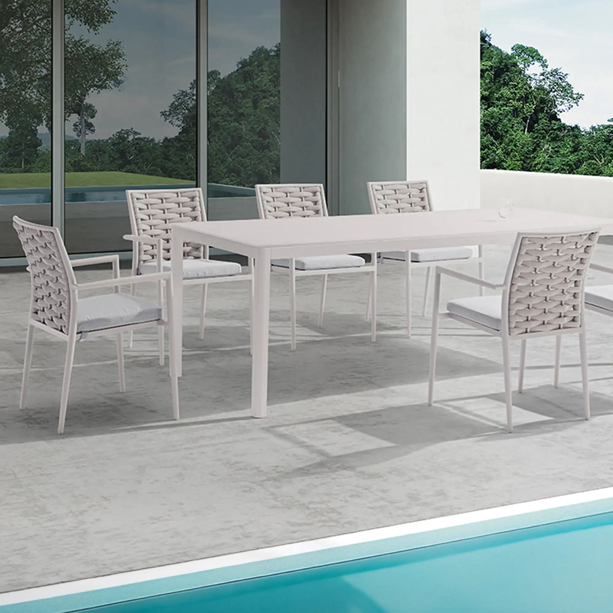 White outdoor dining table and six chairs with woven backs and grey cushions on a patio next to a blue swimming pool, with a modern building and green trees in the background.