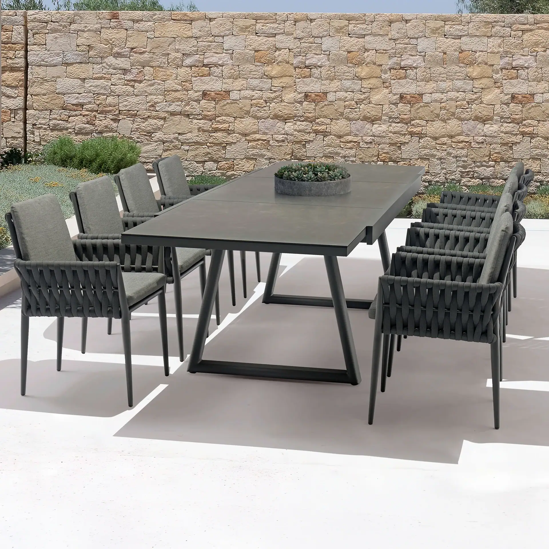 Outdoor dining set with a dark gray table and ten woven chairs on a light patio, backed by a stone wall and green plants.
