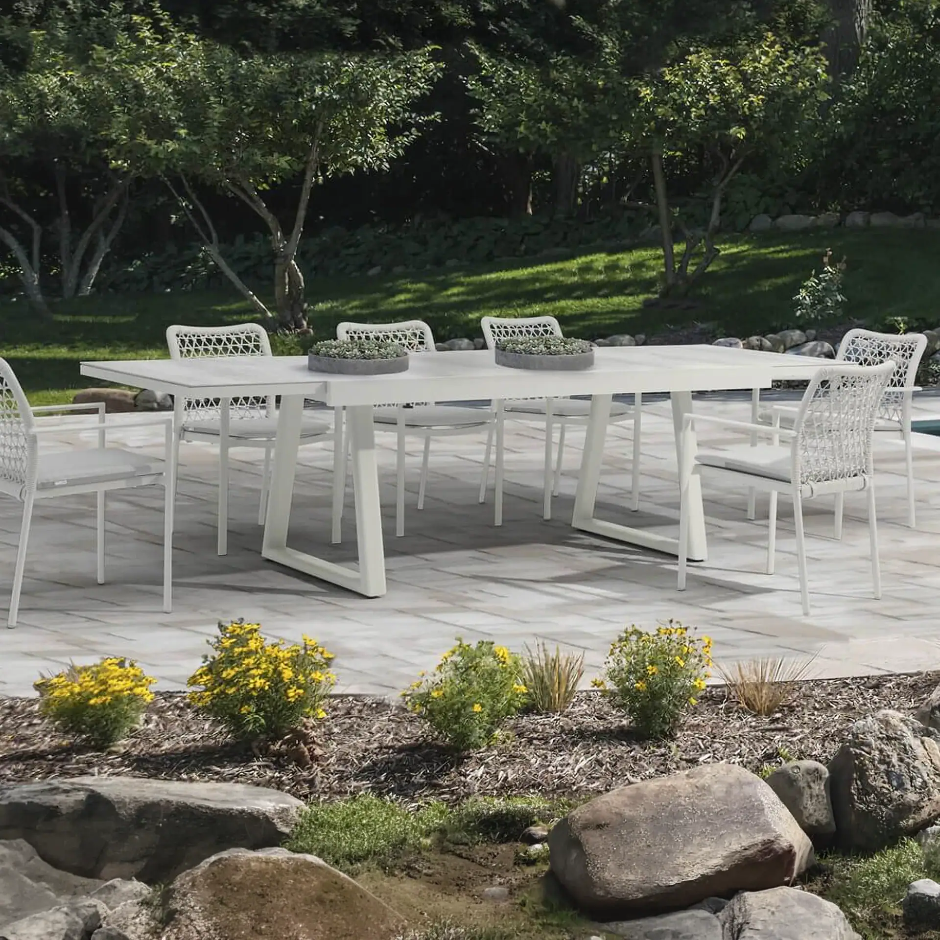 A modern white outdoor dining table with six chairs on a paver patio, surrounded by yellow flowers, rocks, and green trees.
