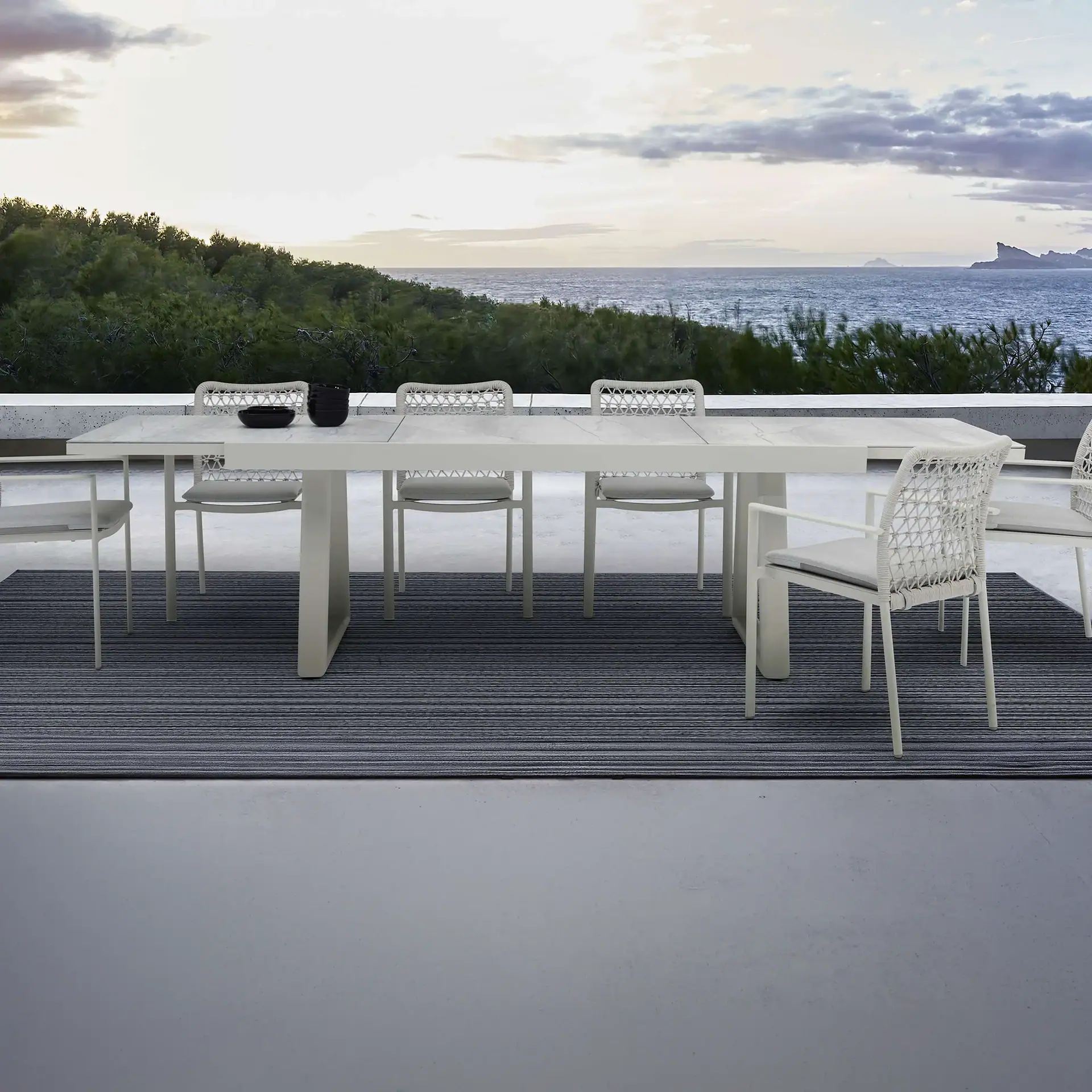 An outdoor dining table and chairs on a patio overlooking a green hillside and the ocean under a cloudy sky.