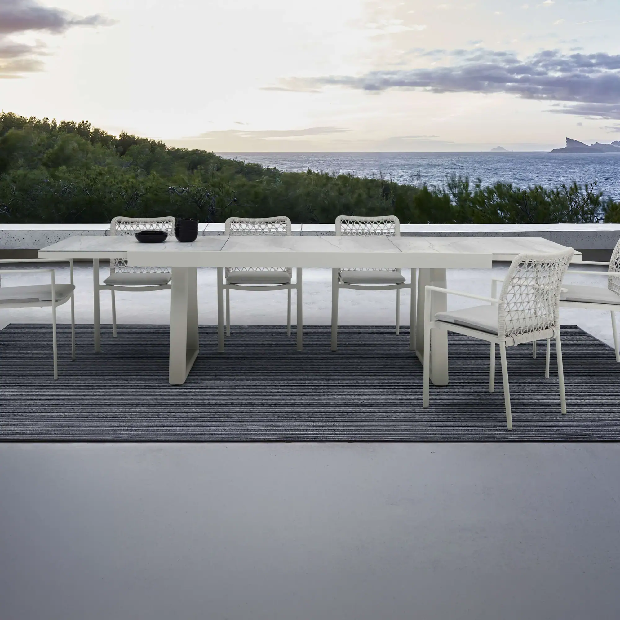 An outdoor dining table and chairs on a patio overlooking a green hillside and the ocean under a cloudy sky.