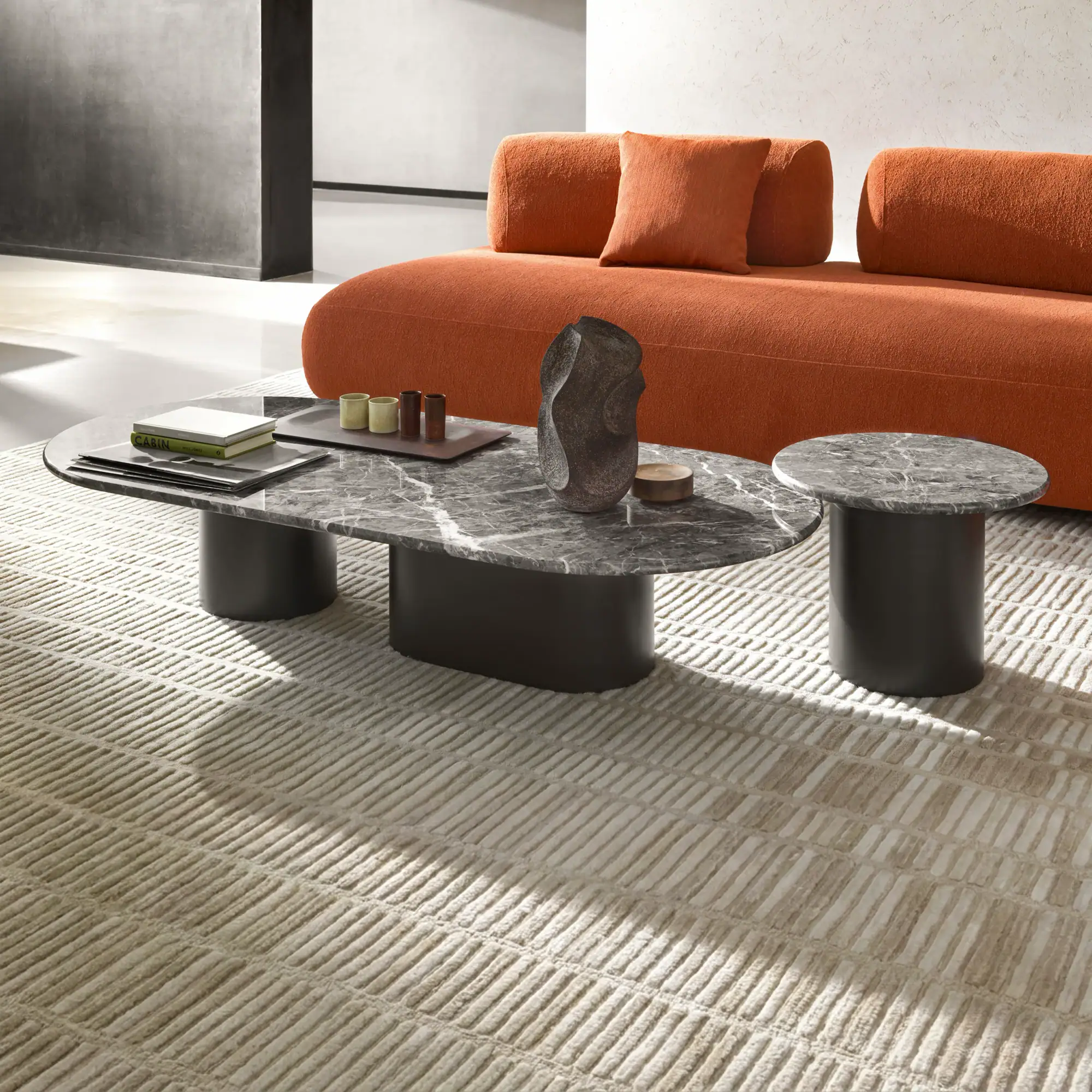 Modern living room with an orange sofa, two gray marble coffee tables, and a patterned beige rug.
