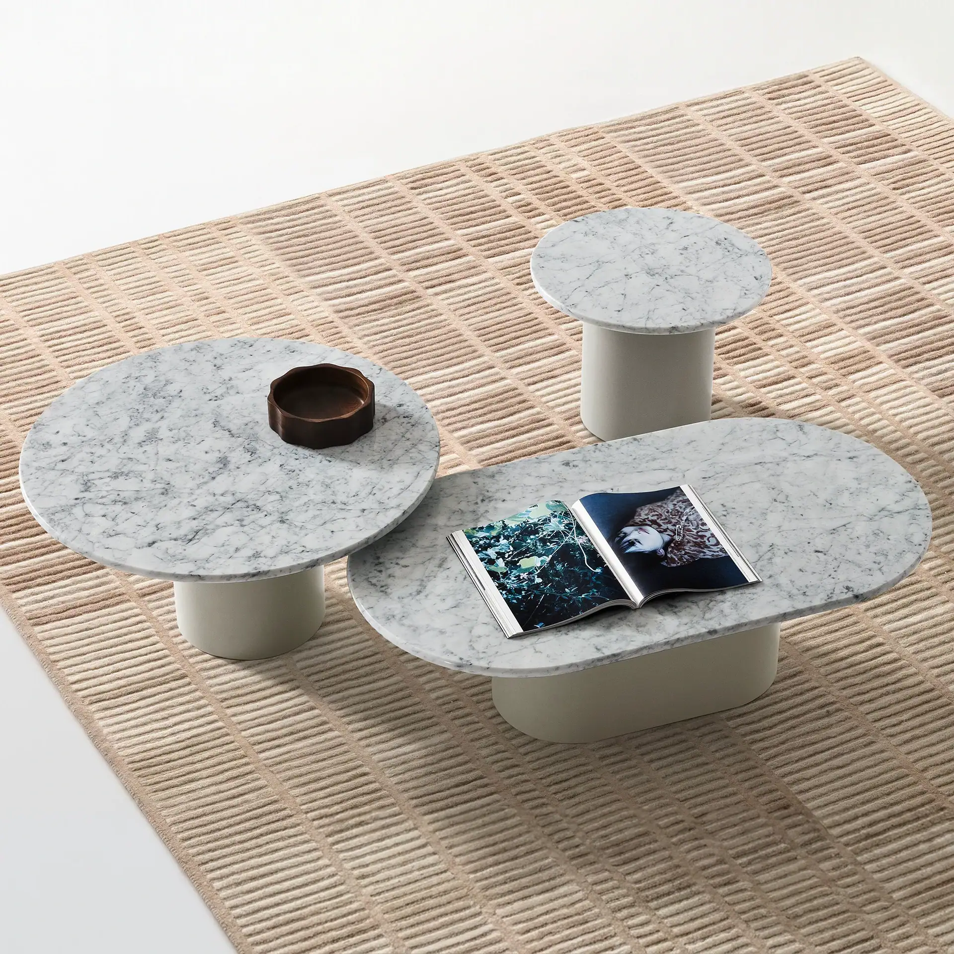 Three marble-topped tables, one oval and two round, on a textured beige rug. An open magazine and a wooden bowl are on the tables.