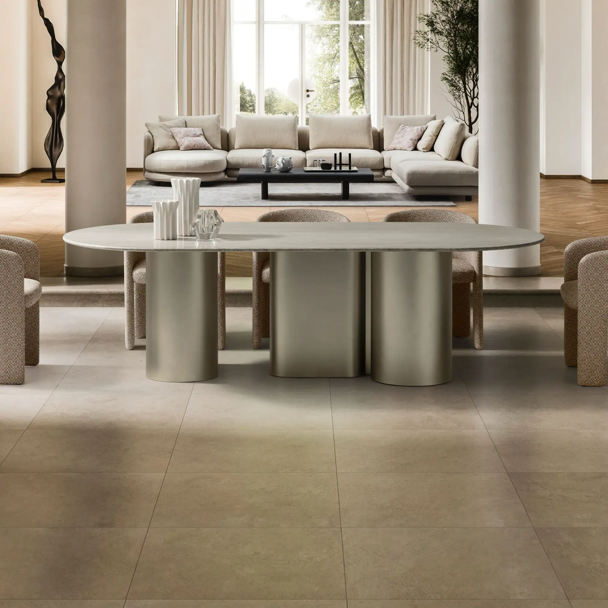 A modern, open-plan living and dining room with an oval dining table, beige sectional sofa, and large window overlooking trees.