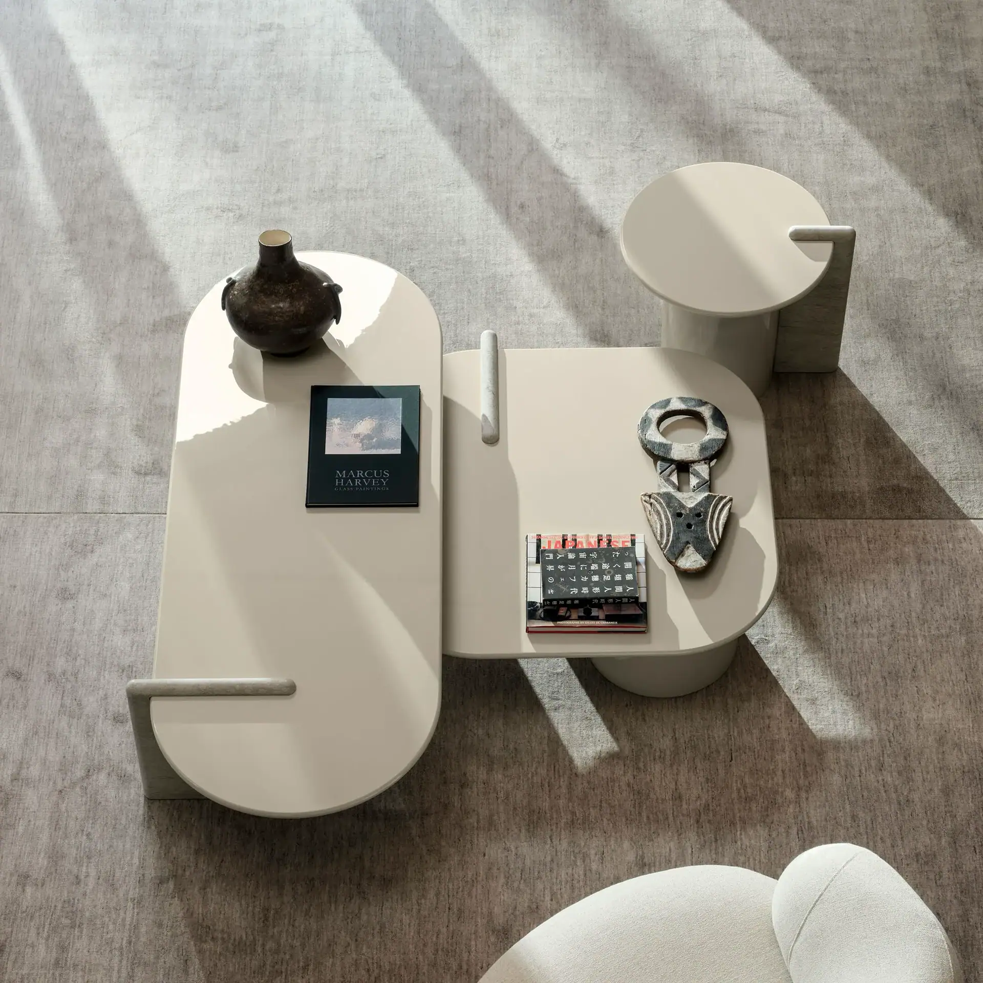 Overhead view of modern beige coffee tables with books and decor on a grey rug, illuminated by diagonal sunlight.