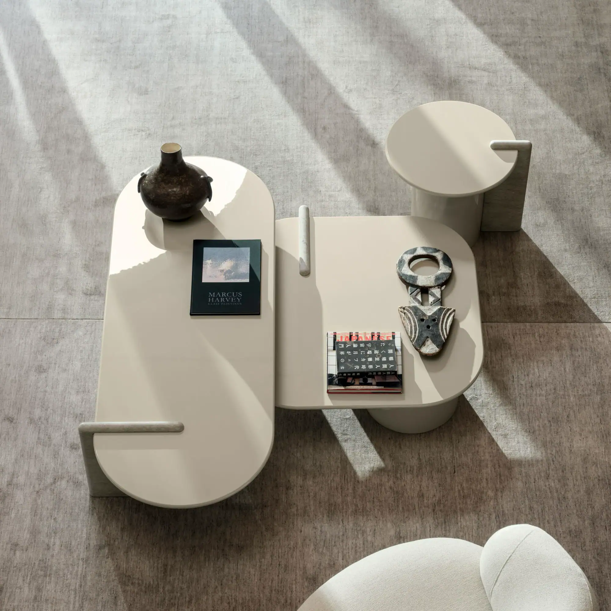 Overhead view of modern beige coffee tables with books and decor on a grey rug, illuminated by diagonal sunlight.